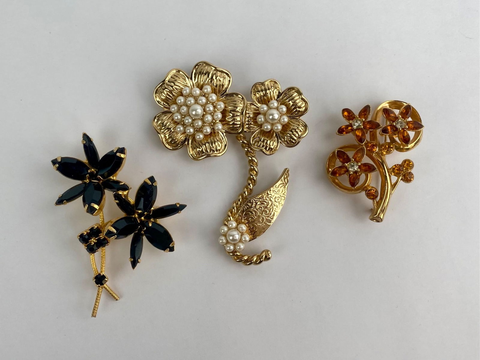 Group of three vintage gold-tone floral brooches featuring rhinestones and faux pearl accents.: Group of three vintage gold-tone floral brooches featuring rhinestones and faux pearl accents. Designs include black marquise-cut rhinestone floral spray, an amber rhinestone scroll-form brooch, and a
