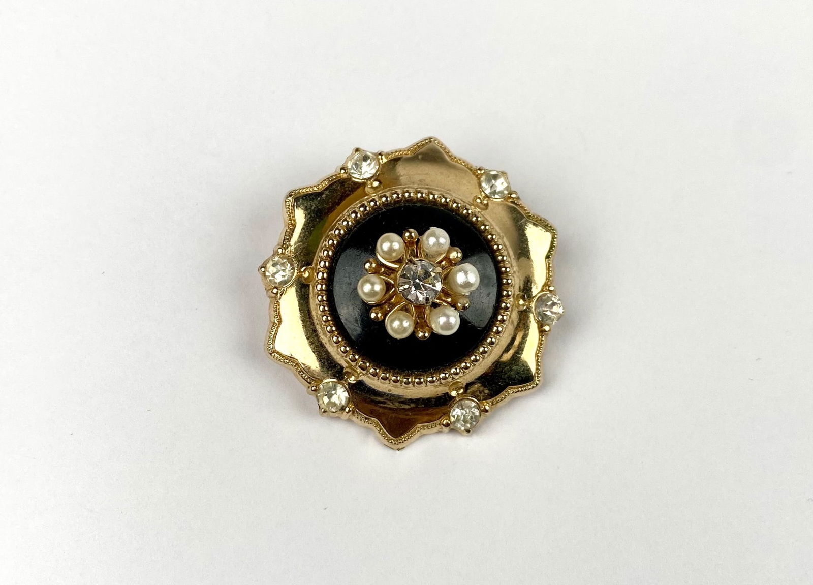 Vintage Coro gold-toned circular brooch featuring a central design of faux pearls surrounding a: Vintage Coro gold-toned circular brooch featuring a central design of faux pearls surrounding a single rhinestone on a black enamel background. The outer edge is adorned with additional rhinestones an