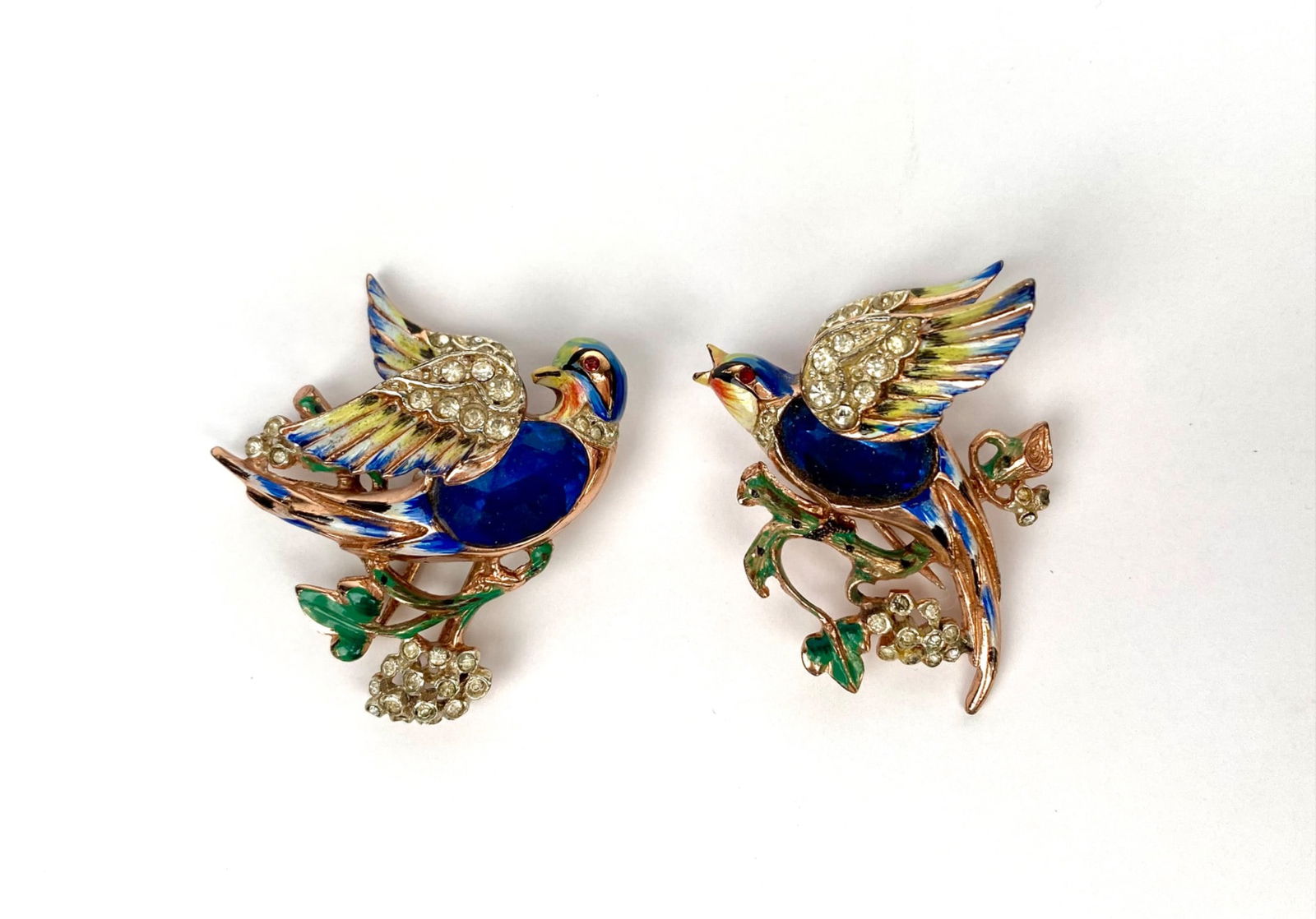 Corocraft Sterling Silver ‘Bill & Coo’ Bird Duette Brooch Set: Corocraft bird-shaped brooches crafted in sterling silver and marked “Sterling” on the reverse. Adorned with colorful enamel detailing in blue, green, and yellow, and accented with rhinestones