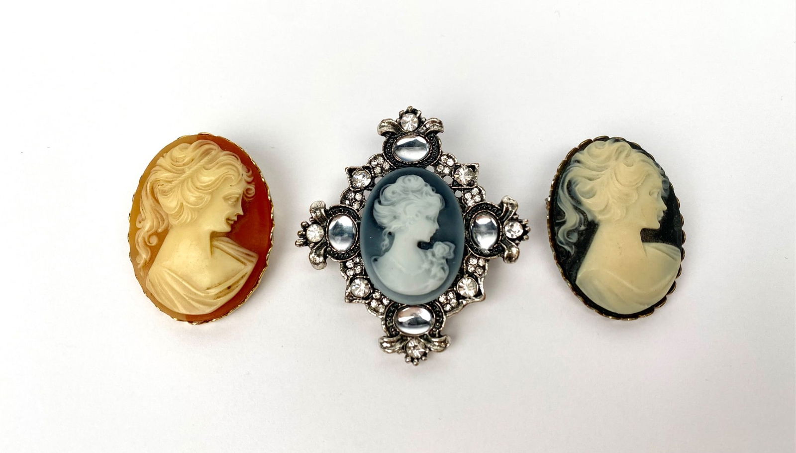 Three vintage cameo brooch pins featuring portrait designs. Includes two oval cameo brooches with: Three vintage cameo brooch pins featuring portrait designs. Includes two oval cameo brooches with relief portraits on contrasting backgrounds and one ornate cross-shaped brooch with an oval cameo cent