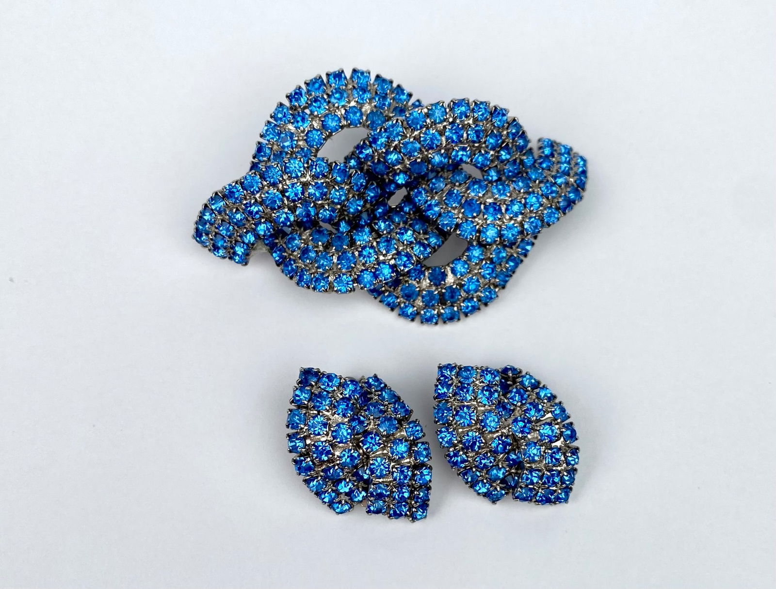 Vintage Blue Rhinestone Knot Brooch & Matching Clip Earrings Set: Vintage brooch and clip earrings set featuring vibrant blue rhinestones. The brooch showcases an intricate knot design with a silver-tone back. The matching clip earrings are marquise-shaped and also