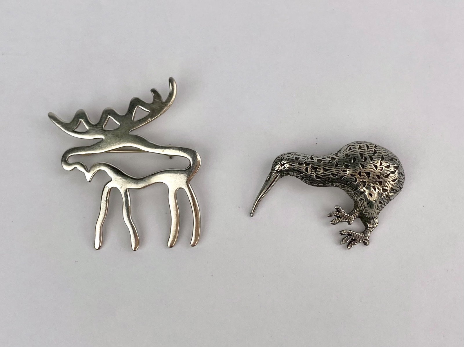 Pair of Silver-Tone Animal Figural Brooches: Pair includes two silver-tone figural animal brooches. One brooch depicts a stylized openwork moose with elongated legs and cutout antler details. The second brooch features a textured kiwi bird with