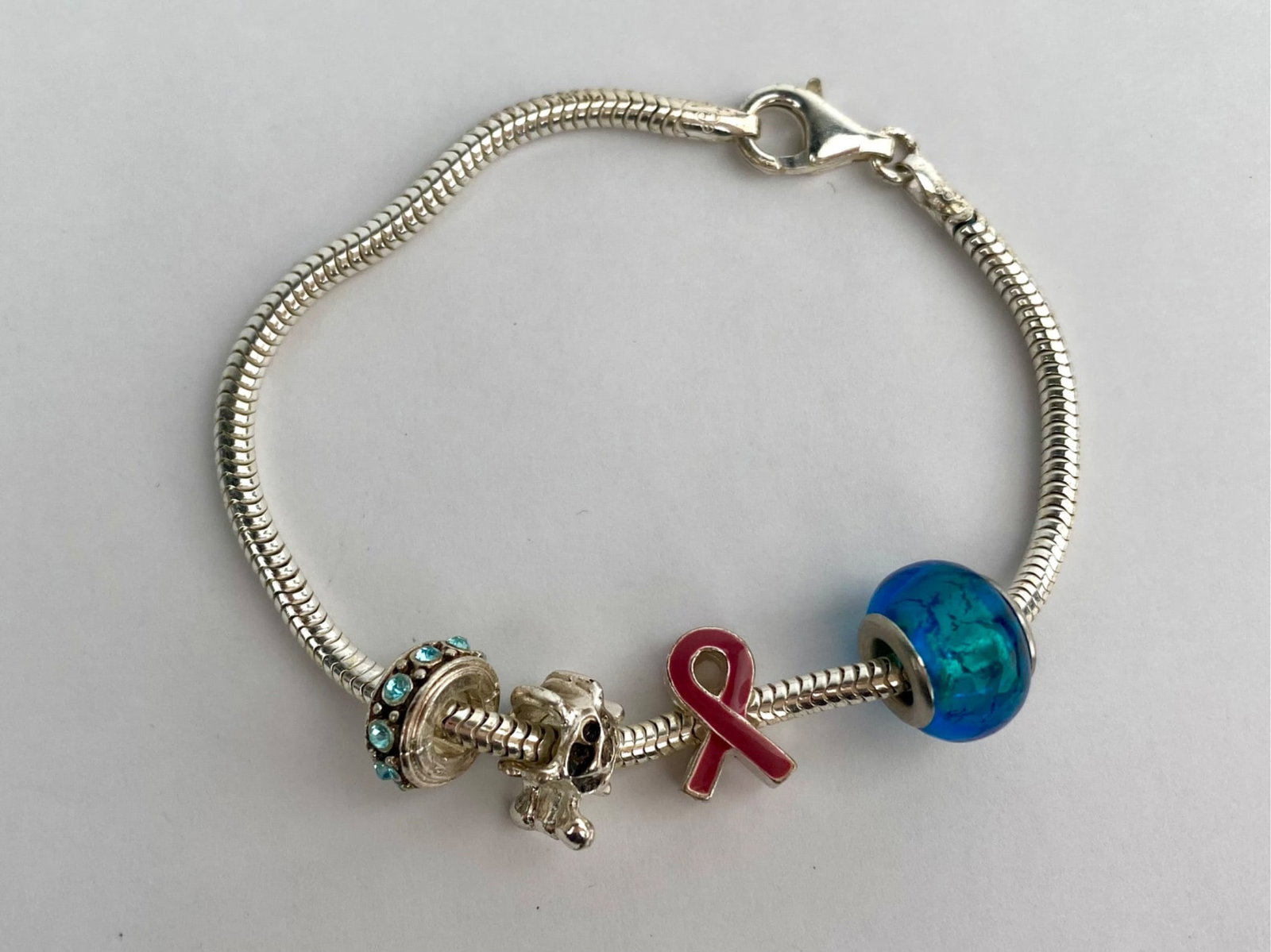 Sterling silver snake chain charm bracelet featuring a lobster clasp closure and several assorted: Sterling silver snake chain charm bracelet featuring a lobster clasp closure and several assorted slide-on charms, including a blue glass bead, an enameled awareness ribbon charm, and a figural turtle
