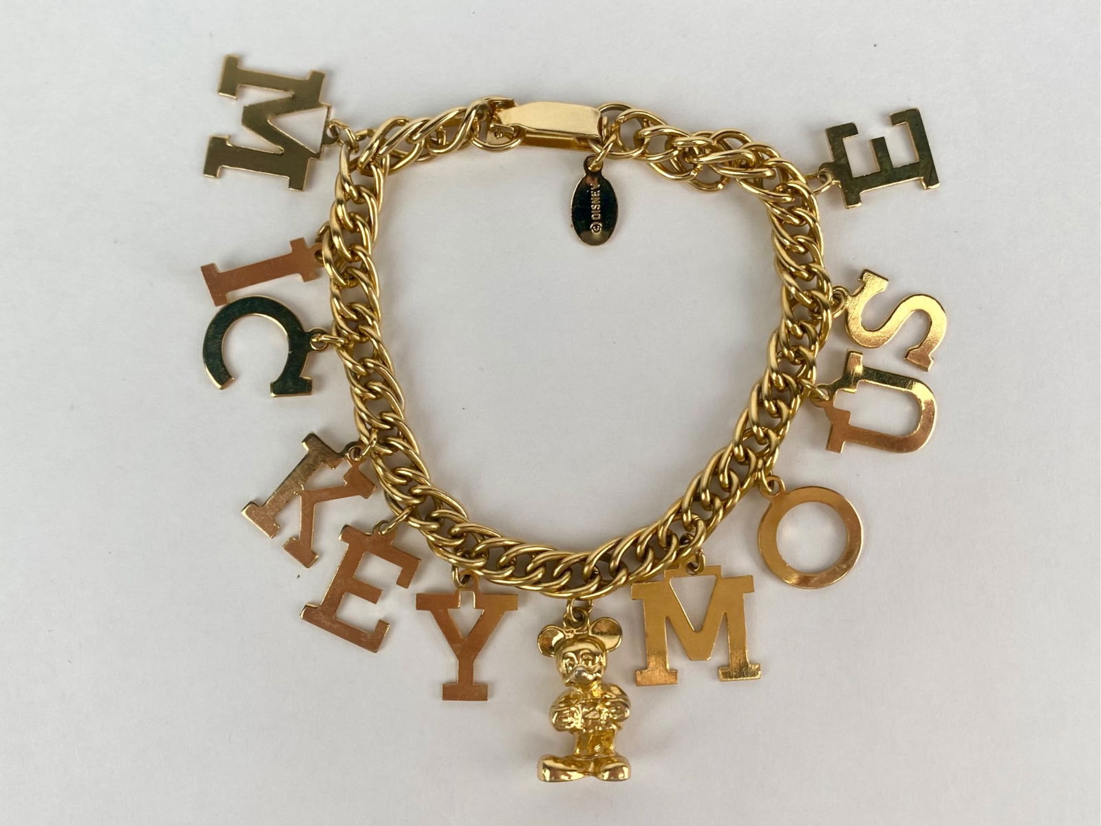 Disney gold-tone charm bracelet featuring block letter charms spelling "MICKEY MOUSE," centered by a: Disney gold-tone charm bracelet featuring block letter charms spelling "MICKEY MOUSE," centered by a dimensional Mickey Mouse figure charm. The bracelet is constructed with a chunky double-link chain