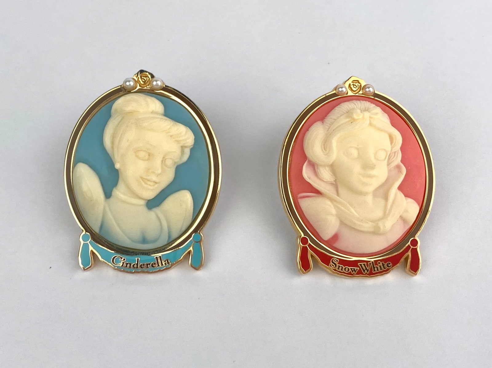 Two-piece Disney pin set featuring cameo-style portrait pins of Cinderella and Snow White, each: Two-piece Disney pin set featuring cameo-style portrait pins of Cinderella and Snow White, each presented in an oval gold-tone frame with molded princess profiles against contrasting-colored backgroun