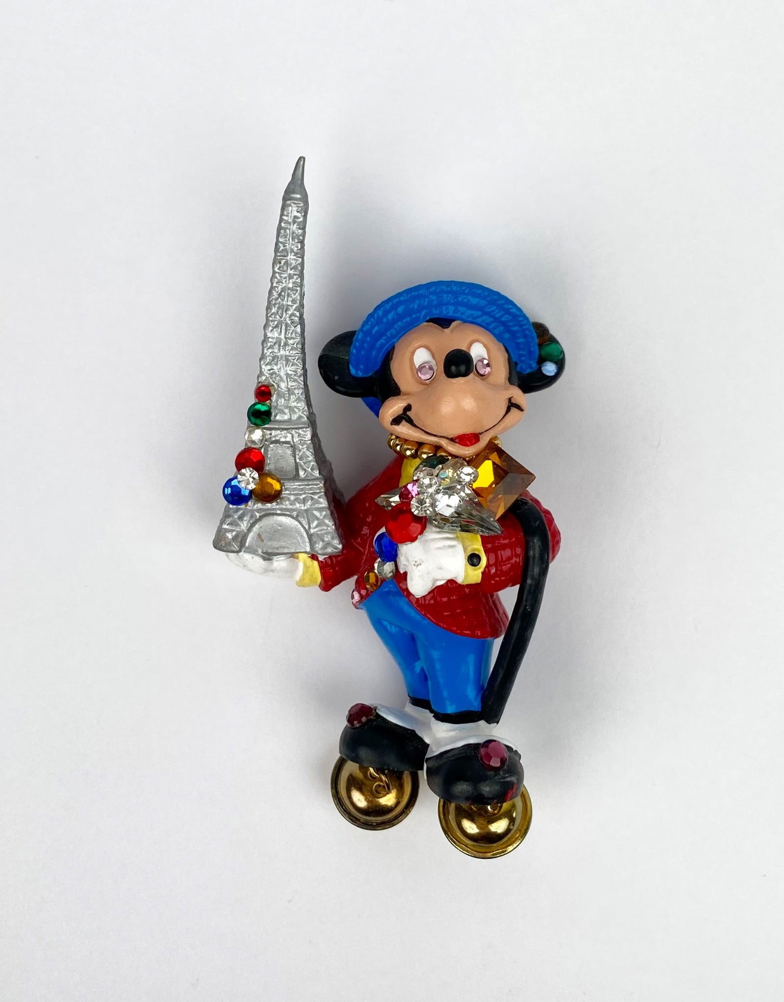 Hand-painted Disney brooch featuring Mickey Mouse holding a silver Eiffel Tower adorned with: Hand-painted Disney brooch featuring Mickey Mouse holding a silver Eiffel Tower adorned with colorful embellishments, produced by Bullyland in Germany. Mickey is dressed in a red jacket, blue pants, a