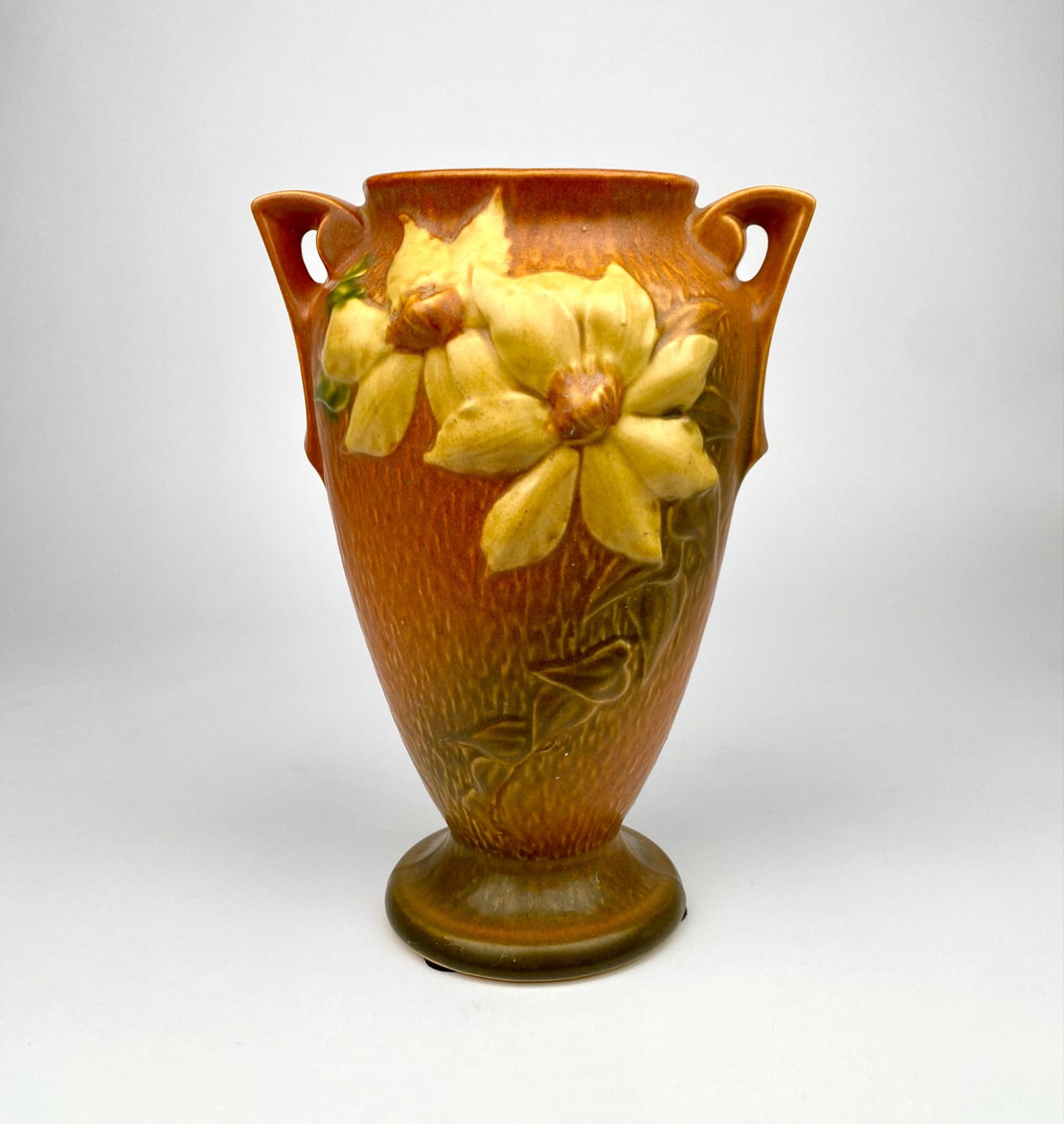 Roseville Pottery Clematis Pattern Vase, Model 111, USA: Roseville Pottery vase in the clematis pattern, featuring a double-handled design with embossed yellow clematis flowers set against an orange and brown textured background. The base is marked ‘Rosev