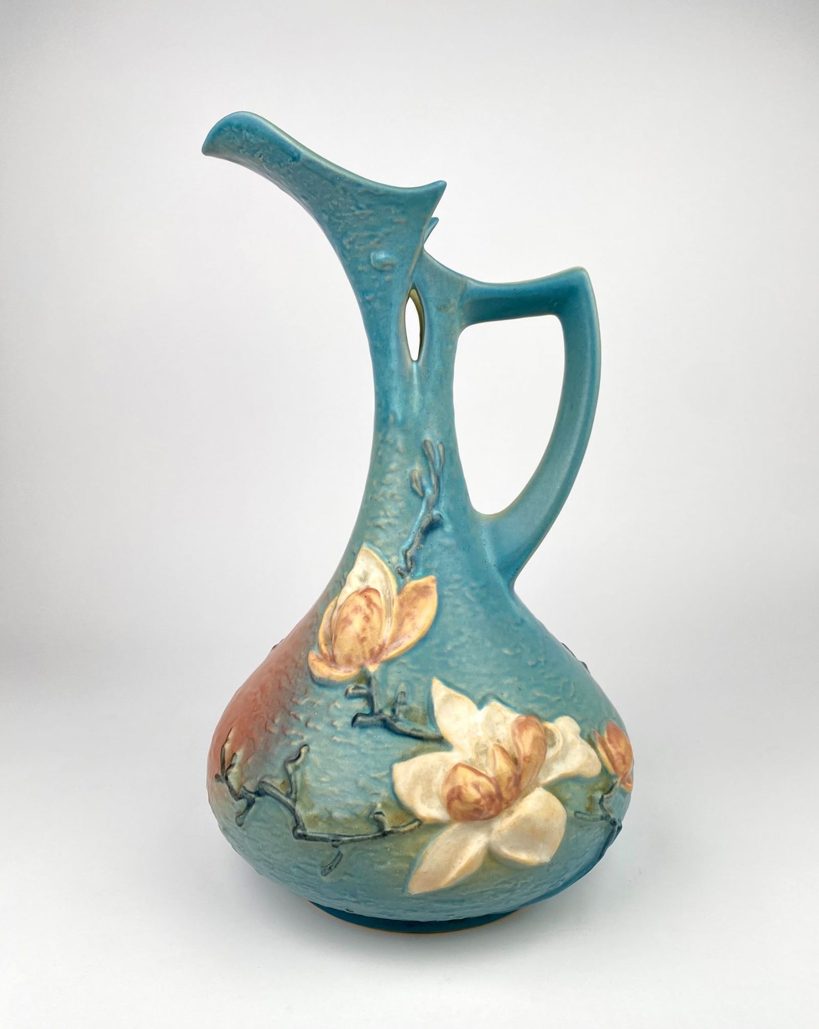 Large Roseville Pottery Blue Magnolia Ewer, Model 15-15, USA: Roseville Pottery Magnolia pattern ewer featuring a light blue gradient base transitioning into soft pink. The ewer is adorned with raised floral reliefs of magnolia blossoms and branches, showcasing