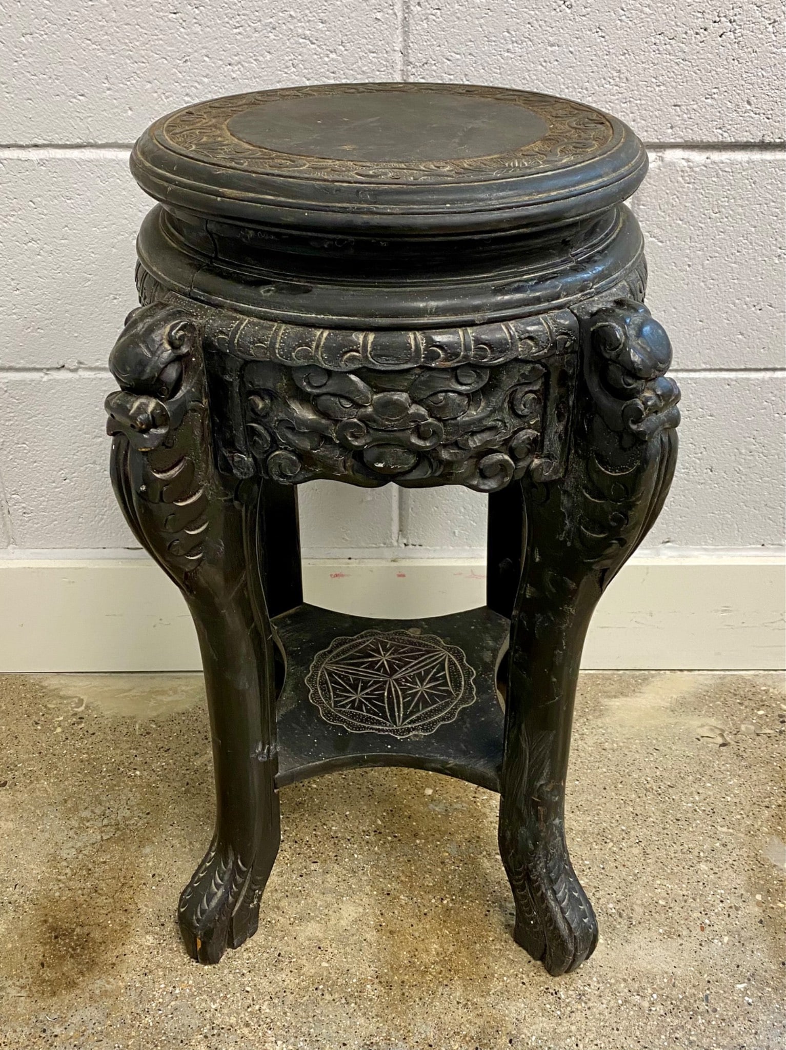 A hand-carved wooden plant stand featuring dragon motif designs on the legs and decorative carvings: A hand-carved wooden plant stand featuring dragon motif designs on the legs and decorative carvings on the apron and round seat. The lower platform bears an etched geometric design. Wear, cracks, and