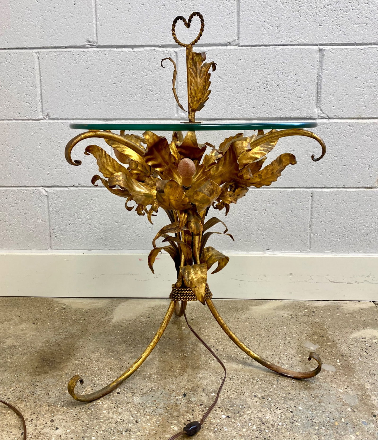 Vintage Hollywood Regency Gilt Tole Floral Lamp Table with Glass Top: Gilded metal side table with a round glass top and integrated lighting. Features floral leaf-shaped metalwork detailing and three-light fixtures. Includes a heart-shaped metal topper. Wiring and