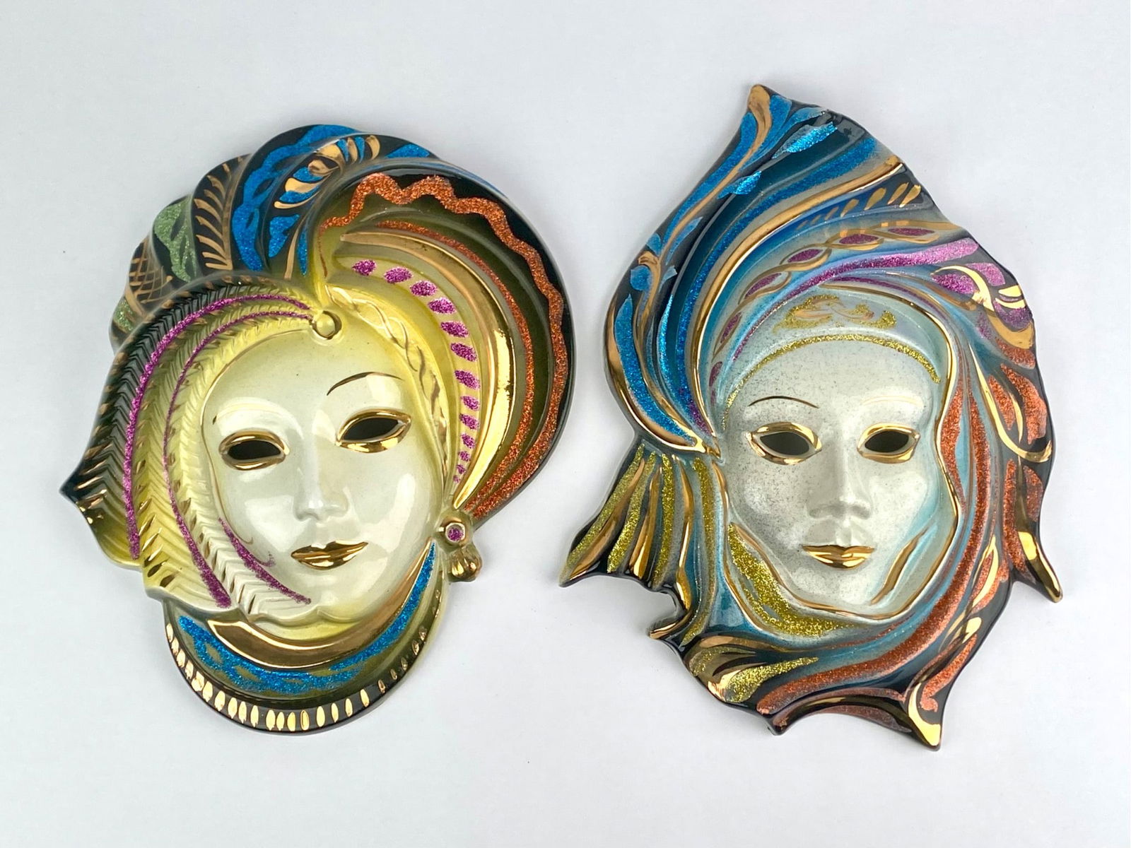 Lot includes a pair of decorative Venetian-style ceramic wall masks. Each mask depicts a stylized: Lot includes a pair of decorative Venetian-style ceramic wall masks. Each mask depicts a stylized face wearing an elaborate headdress highlighted with multicolored metallic accents and glitter decorat