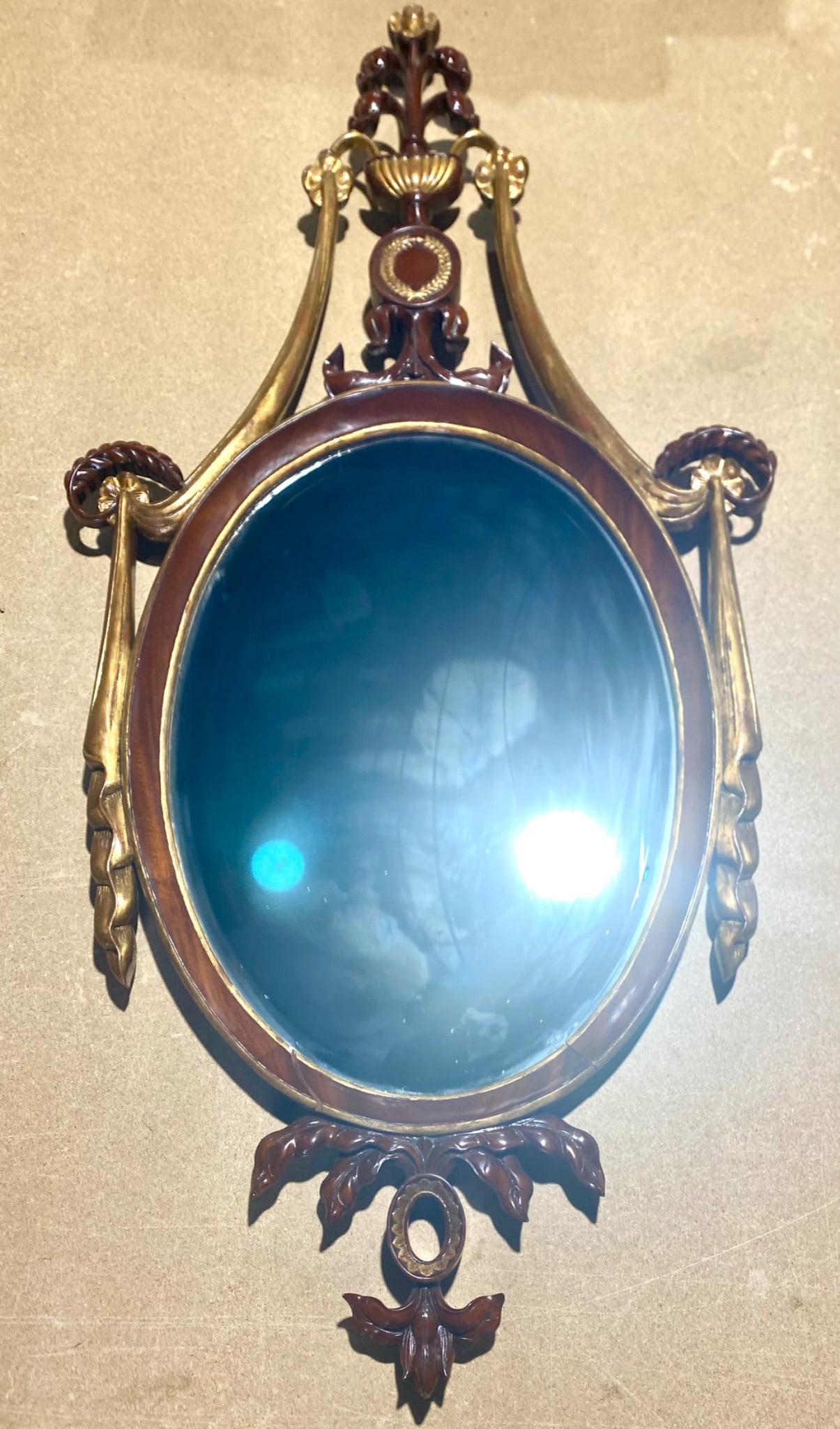 Decorative wall mirror featuring an oval glass plate within a carved wood frame accented with gilt: Decorative wall mirror featuring an oval glass plate within a carved wood frame accented with gilt decoration. The frame displays foliate and scroll motifs with an openwork crest and pendant detail. C