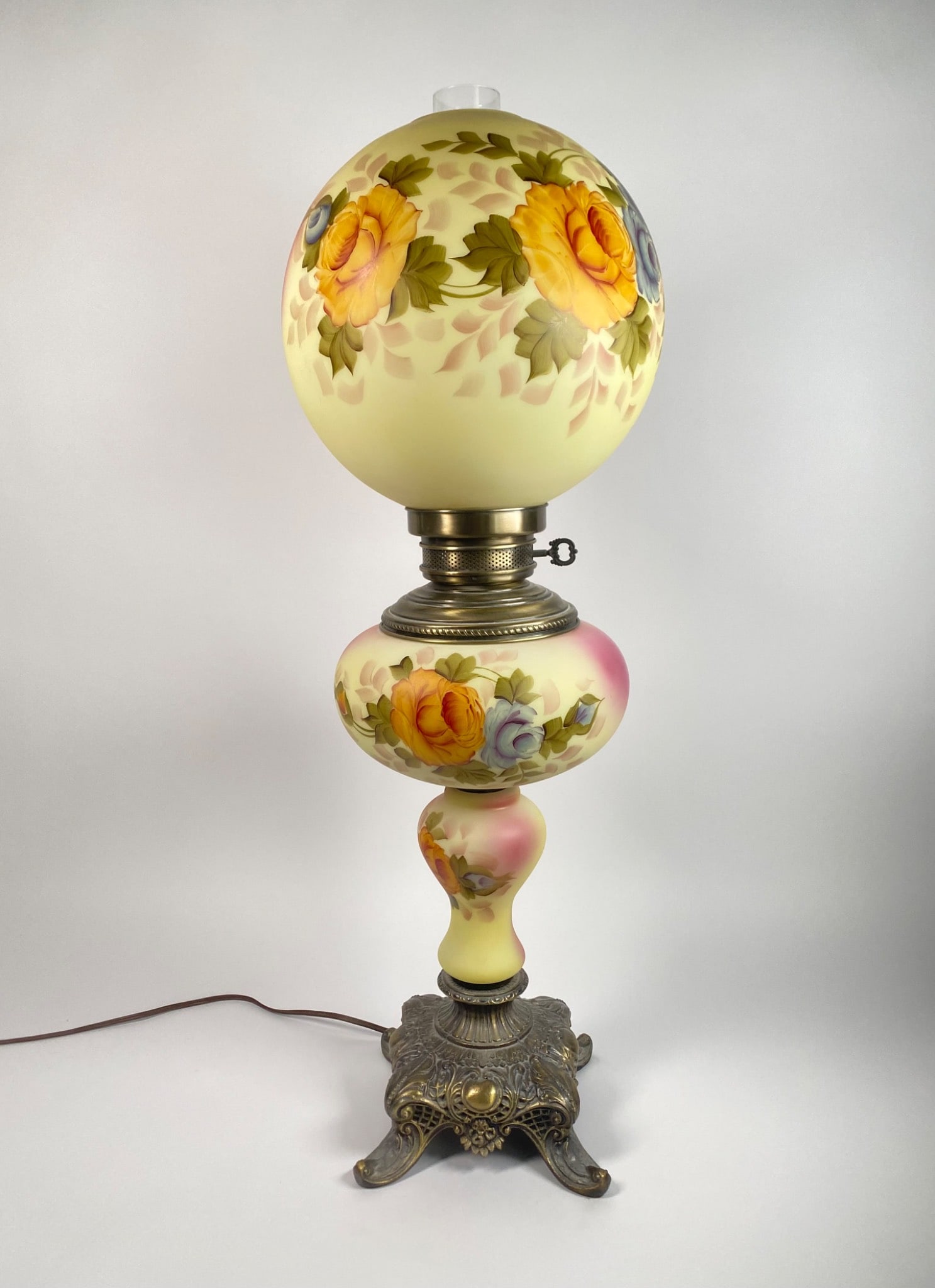 Hand-painted floral glass hurricane lamp featuring vibrant yellow and blue roses with green foliage.: Hand-painted floral glass hurricane lamp featuring vibrant yellow and blue roses with green foliage. The lamp includes a frosted and decorated glass shade and body, resting on a detailed brass base wi