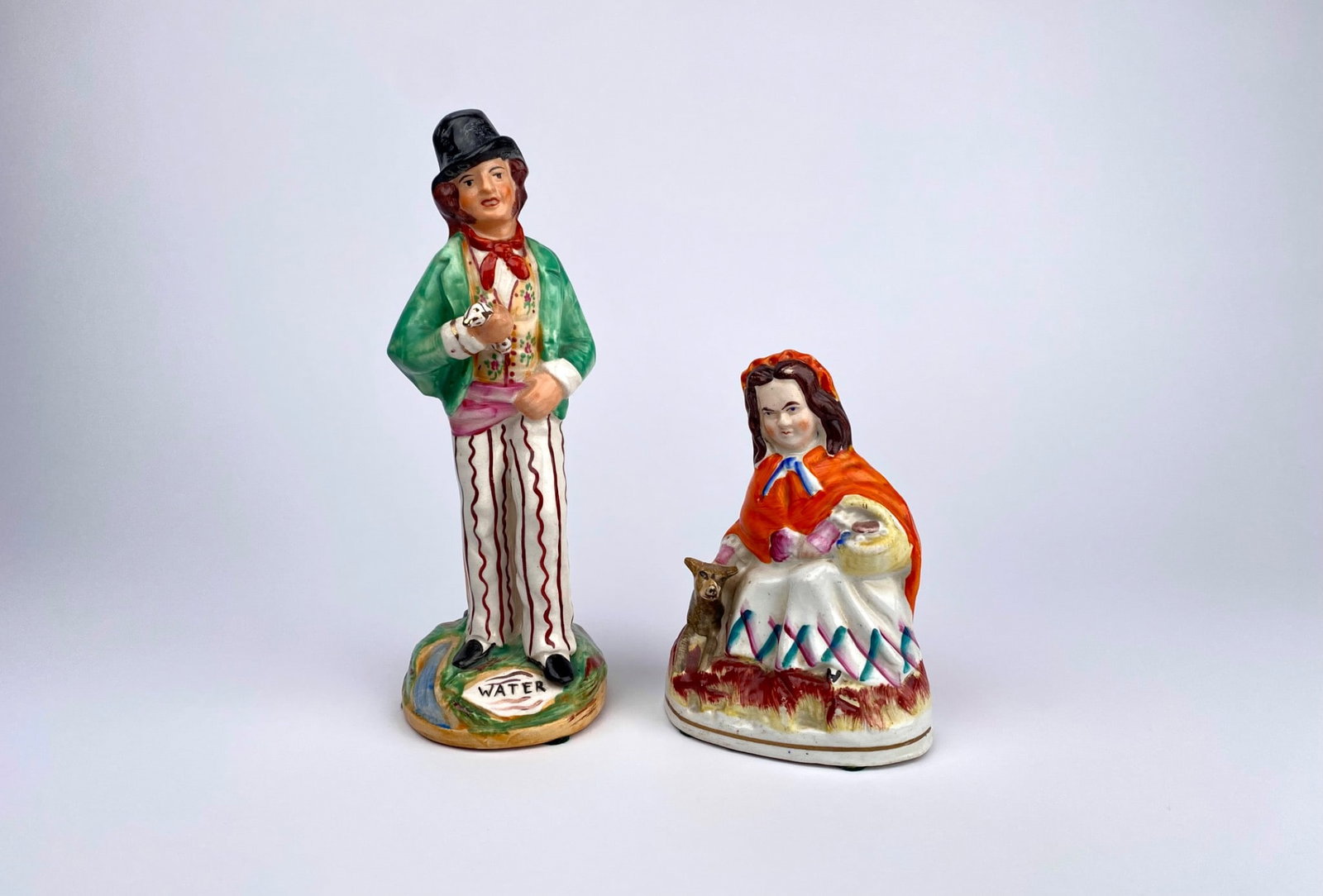 Antique English Staffordshire Figurines: Man with ‘Gin/Water’ Theme & Woman with Dog: Staffordshire figurines, including a man in a green coat and striped pants with ‘Gin/Water’ inscriptions on the base, and a woman in an orange shawl with a dog by her side. Each figure is