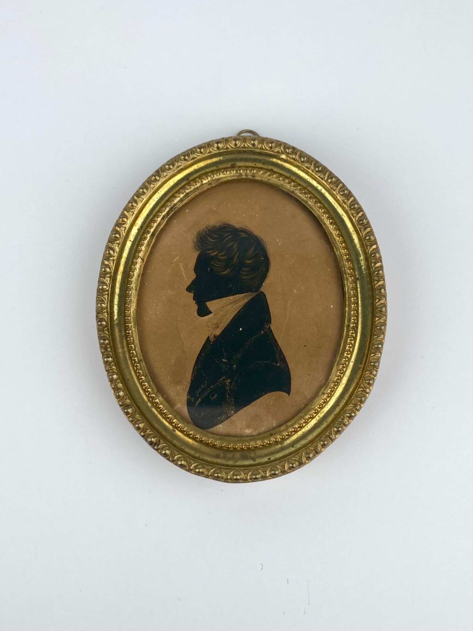 Early 19th century silhouette portrait depicting a gentleman in profile, executed in black against a: Early 19th century silhouette portrait depicting a gentleman in profile, executed in black against a warm tan background. The silhouette is housed in a gilt metal oval frame with beaded and foliate mo