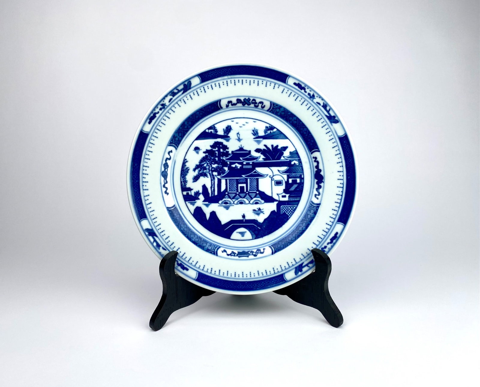 Chinese blue and white porcelain decorative plate featuring a traditional Chinese-inspired landscape: Chinese blue and white porcelain decorative plate featuring a traditional Chinese-inspired landscape design, including pagodas, trees, and bridges. Marked ‘Made in China’ on the back with addition