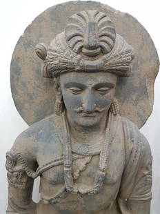 SUBSTANCIAL GANDHARA OF BUDDHA BODHISATTVA - 32": Ca.300AD. A substantial youthful and benign grey schist statue of the bodhisattva Avalokiteshvara on a plinth, seated wearing a dhoti and sandals with swags of beads and braided ornaments to the neck,