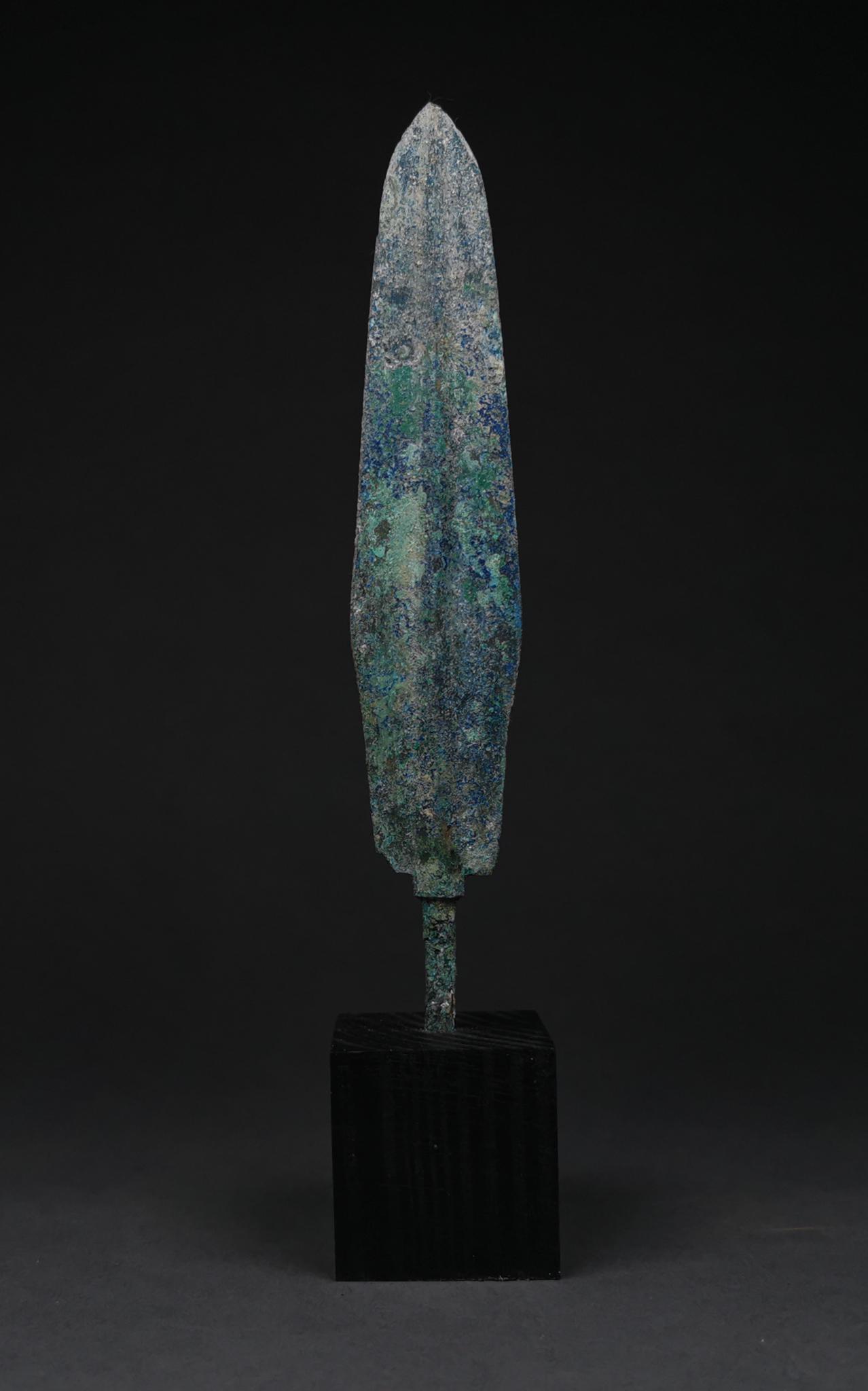 ANCIENT BRONZE SPEAR HEAD ON STAND: 1200-800 BC, Greek Archaic Period. A bronze spearhead with a lozenge-shaped, lentoid sectioned blade, raised midrib and a short tang. Bronze weaponry production flourished in western Asia, the Aegean,