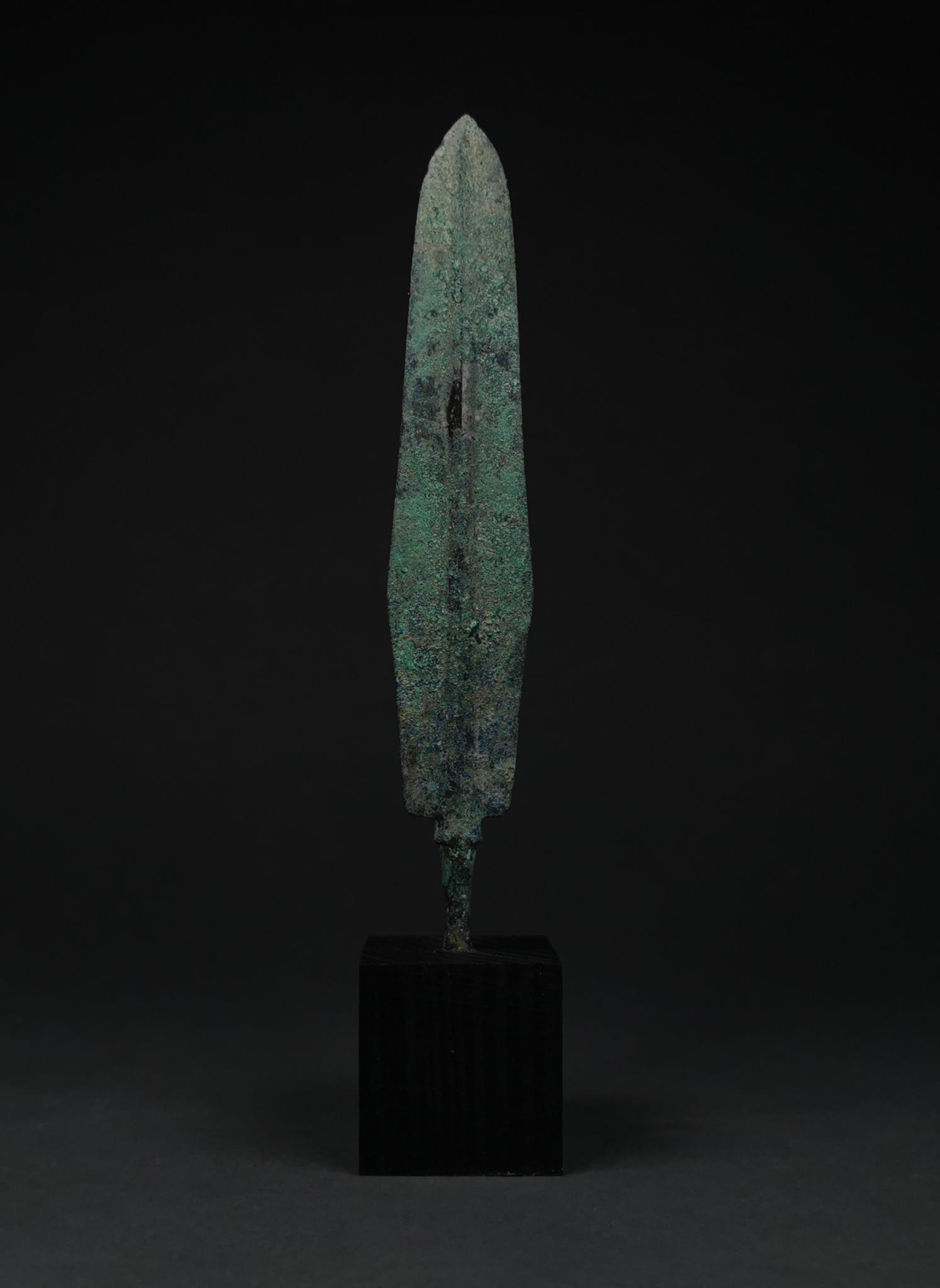 ANCIENT BRONZE SPEAR HEAD ON STAND: 1200-800 BC, Greek Archaic Period. A bronze spearhead with a lozenge-shaped,lentoid sectioned blade, raised midrib and a long tang. This item is mounted on a stand. Bronze weaponry production flouri