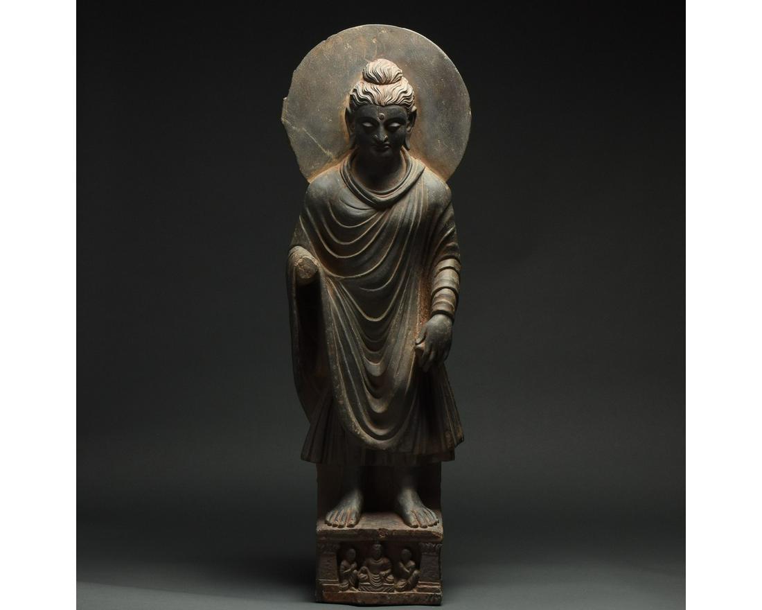 HUGE GANDHARA SCHIST STONE FIGURE OF BUDDHA WITH HALO -: 100 – 300 AD. Gandharan. A stunning schist figure of a standing buddha figure. This excellent figure is backed by a halo and dressed in a flowing Kasaya (monastic robe). The head features thick, ric