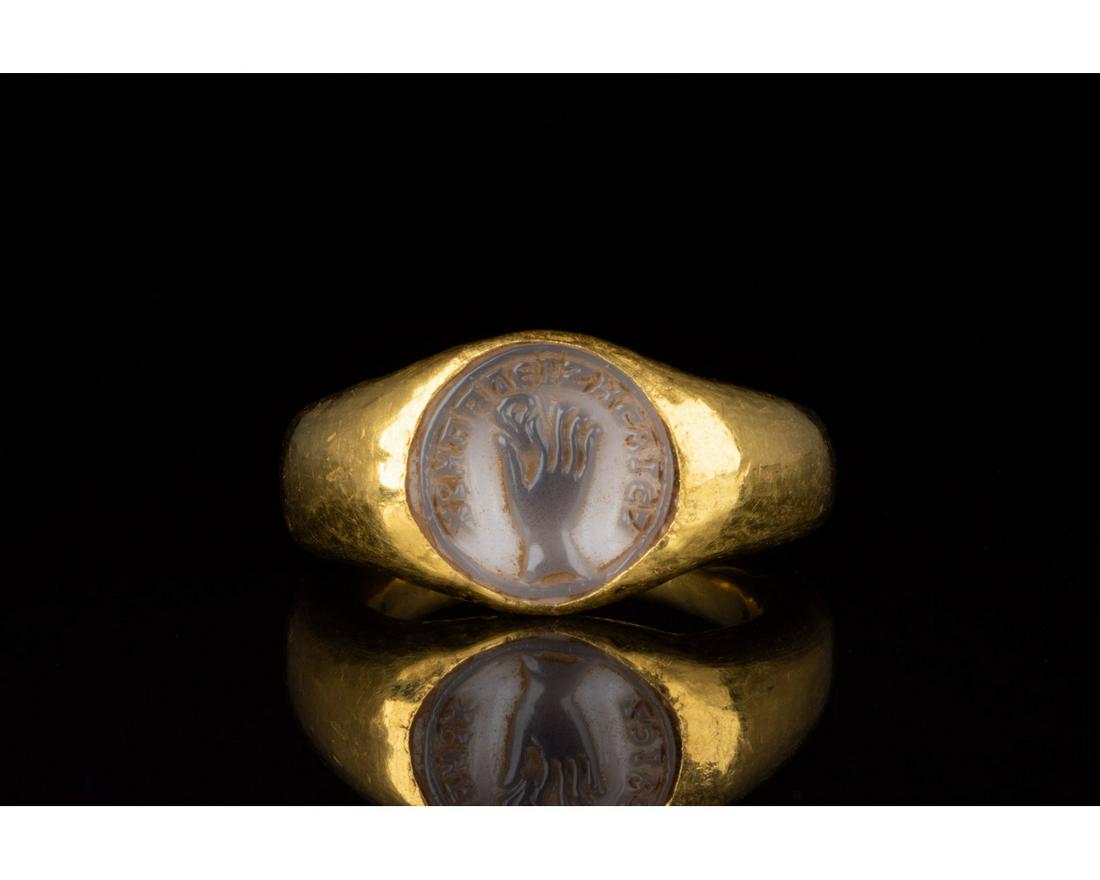 RARE ROMAN GOLD CAMEO RING WITH HAND AND SCRIPT: 100-300 AD. Roman. Gold ring with circular hoop and rounded bezel containing a grey gemstone intaglio setting. The intaglio features a hand pinching an ear, surrounding by a Greek formula requesting t