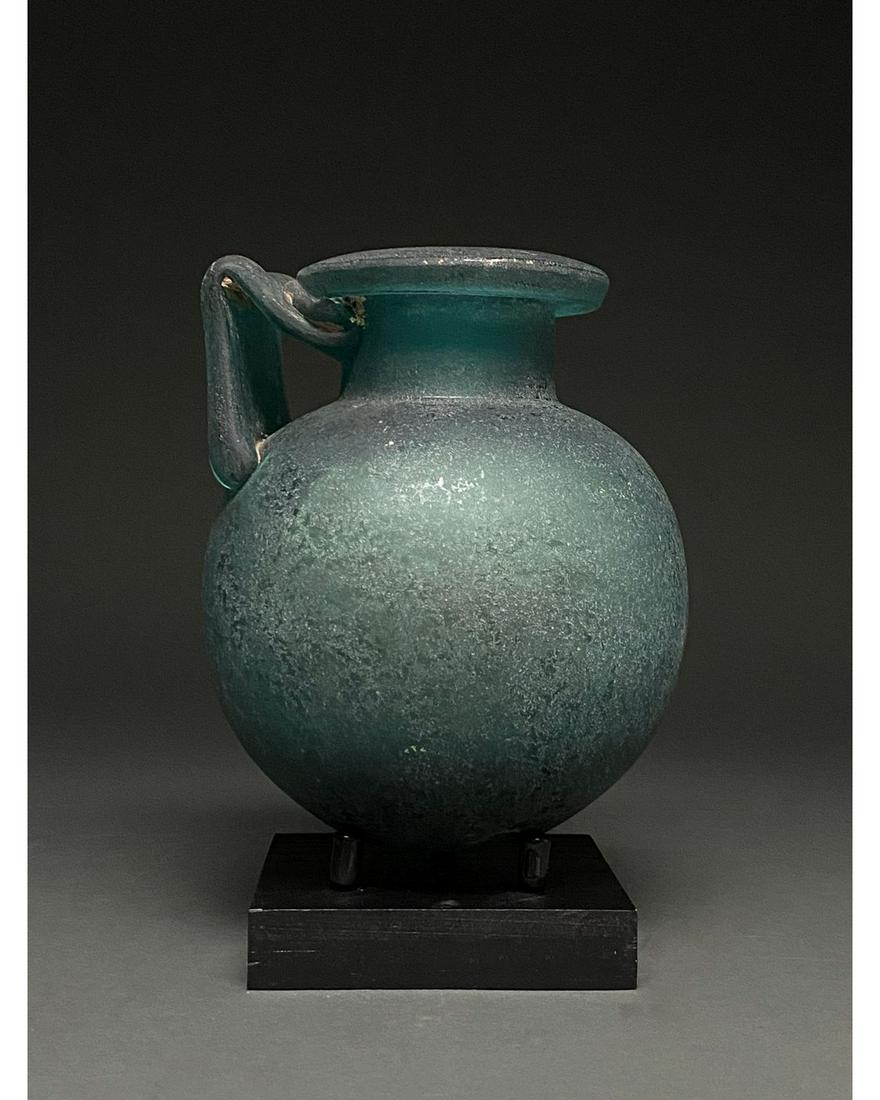 ROMAN STYLE GLASS FLASK: Possibly Grand Tour period. Roman-style greenish-blue glass vessel with an out-turned rim, cylindrical neck, globular body and L-shaped handle. Glass was a major manufacturing industry in the Roman Em