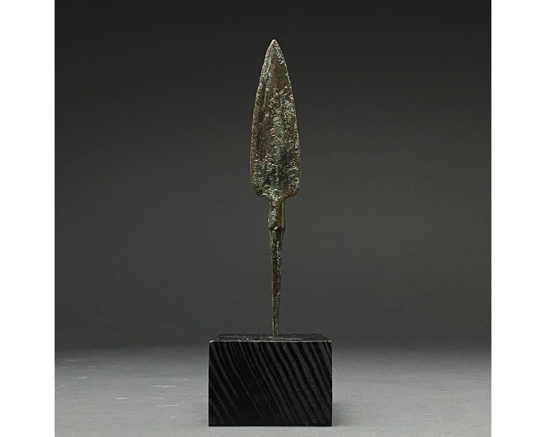 ANCIENT BRONZE SPEAR HEAD ON STAND: 1200-700 BC, Greek Archaic Period. Bronze Age. A bronze spearhead with a leaf-shaped, lentoid sectioned blade, a wide raised midrib and a short neck that transitions into a tang with a pointed termina