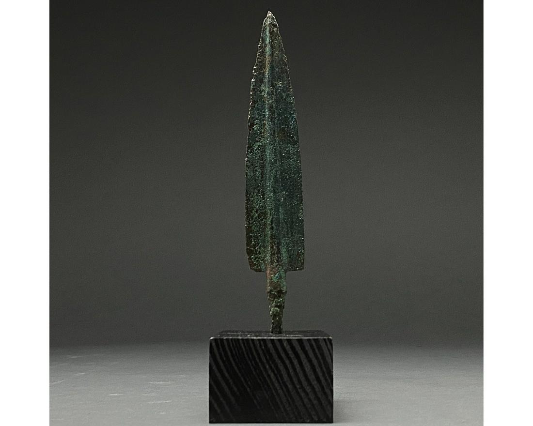 ANCIENT BRONZE SPEAR HEAD ON STAND: 1200-700 BC, Greek Archaic Period. Bronze Age. A bronze spearhead with a leaf-shaped, lentoid sectioned blade, a wide raised midrib and a short neck that transitions into a small tang with a pointed t