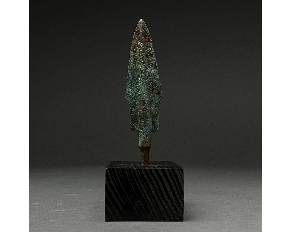 ANCIENT BRONZE SPEAR HEAD ON STAND: 1200-700 BC, Greek Archaic Period. Bronze Age. A bronze spearhead with a leaf-shaped, lentoid sectioned blade, a raised midrib and a short neck that transitions into a small tang with a pointed termin