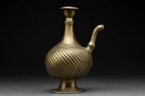 INDIAN MUGHAL OR DECCAN BRASS EWER: Ca. 16th century AD A brass ewer rising from a wide conical-shaped foot with a frilled edge through a drop-shaped body with diagonal ribs and tapering neck, pronounced collar, sloped rim, and curved s