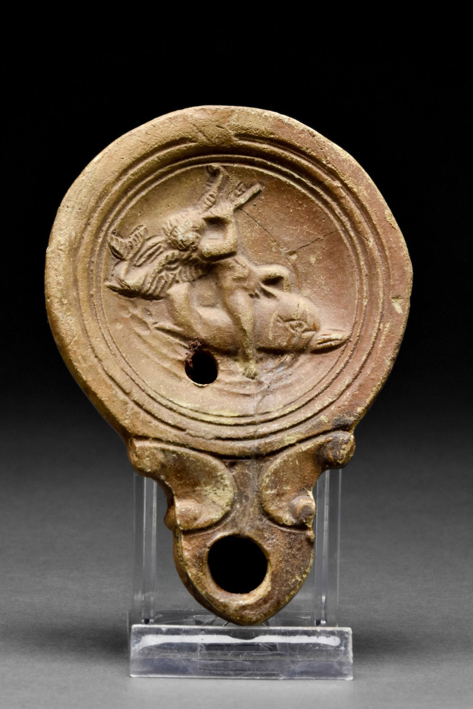 ROMAN TERRACOTTA OIL LAMP WITH EROS RIDING ON A DOLPHIN: Ca. 1-200 AD A flat-bottomed terracotta oil lamp of Loeschcke type VIII, shoulder VIIIb. On the discus low relief figure of Eros playing the flute while riding on a dolphin.Size: L:100mm / W:70mm ; 75