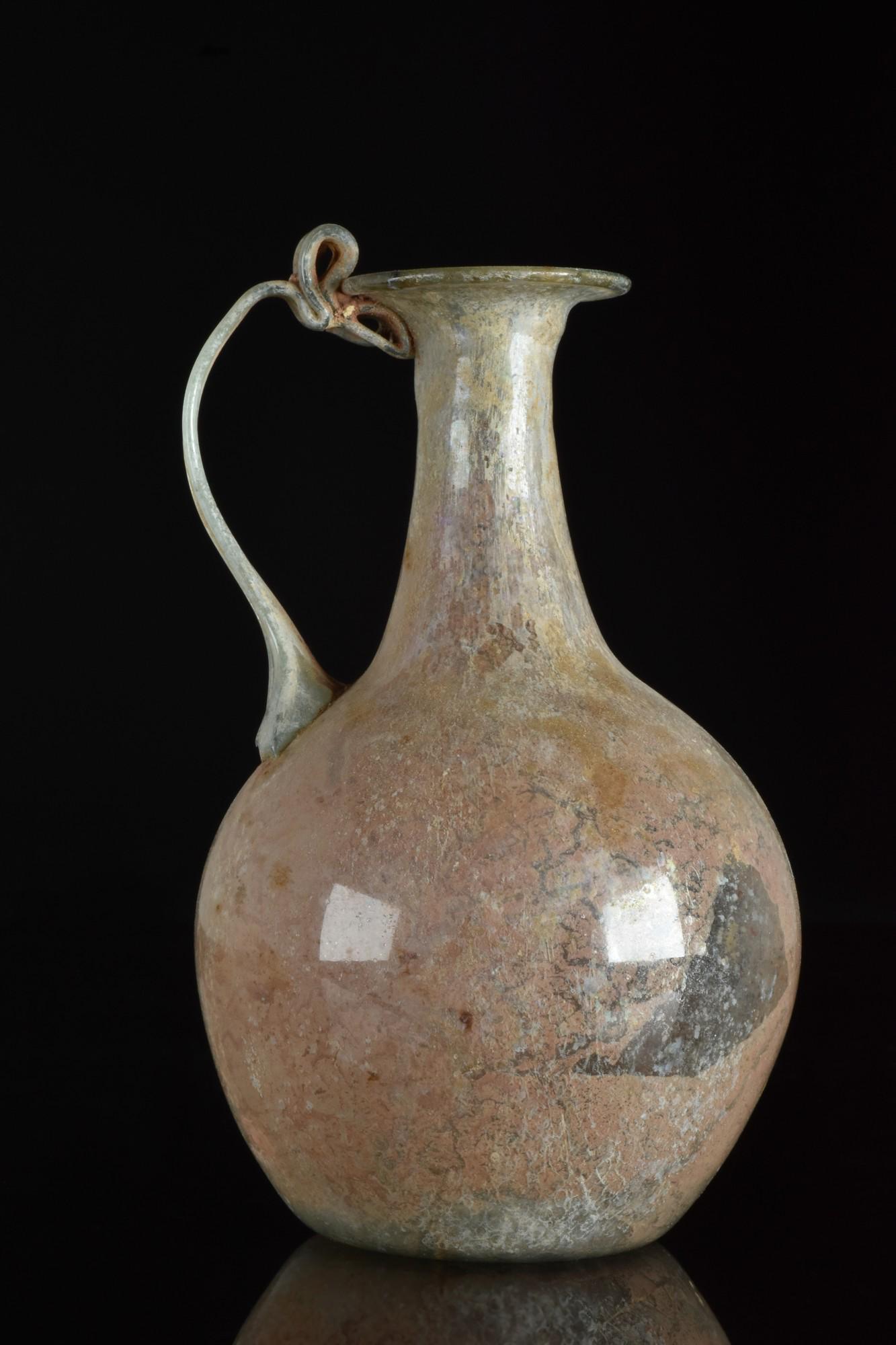 ROMAN LIGHT BLUE-GREEN GLASS JUGLET: Eastern Mediterranean, Ca. 1-100 AD A light clear bluish-green color glass juglet, having a thin ribbon type handle with a loop rigaree at the top. Slightly tapered bulbous body. Overall interior surf