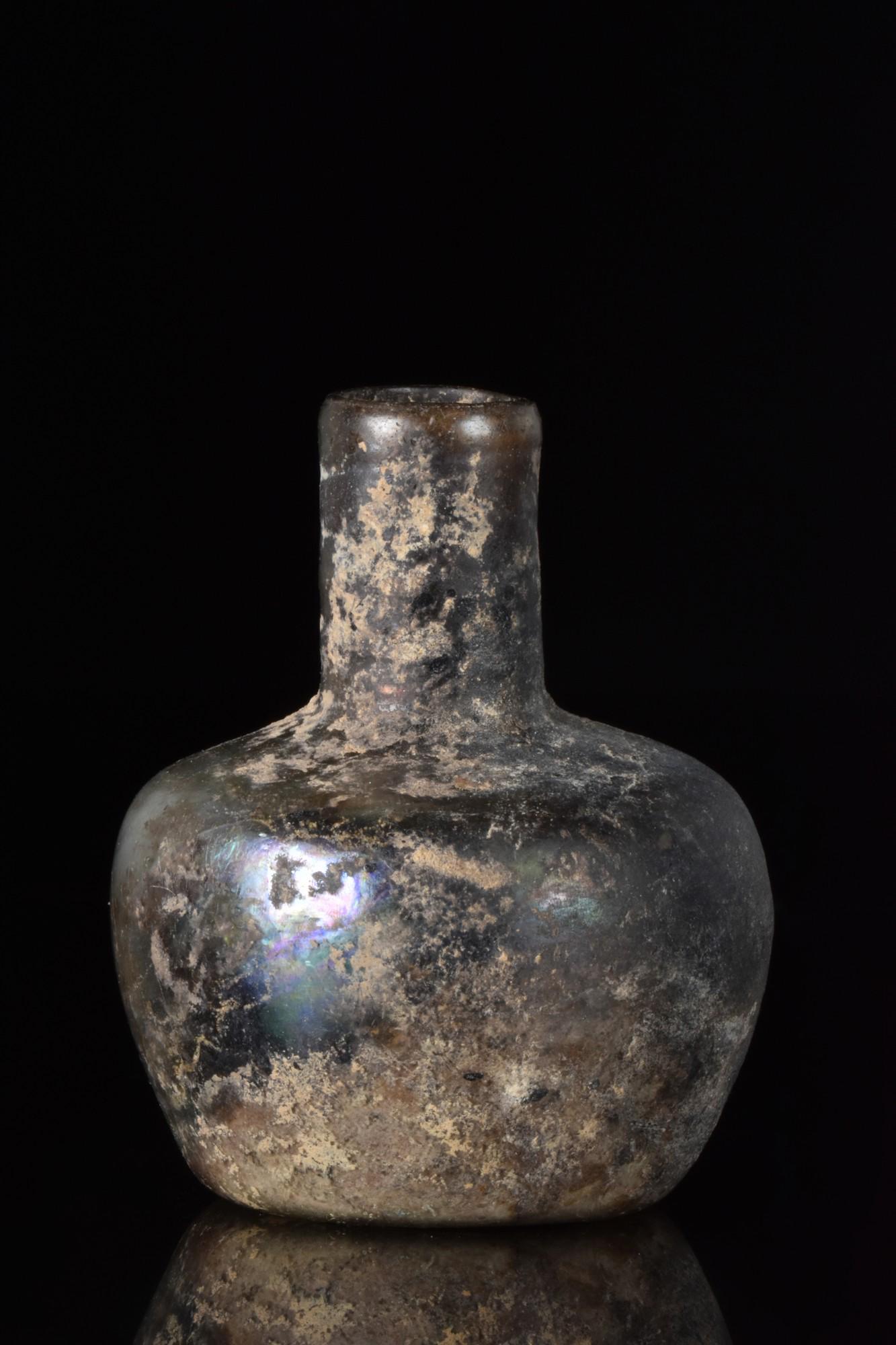 ROMAN GLASS FLASK: Ca. 100-200 AD A glass flask with a beautiful iridescence with a tubular neck and bulbous-shaped lower body with broad shoulder and a concave base. Some weathering and incrustation occur throughout th