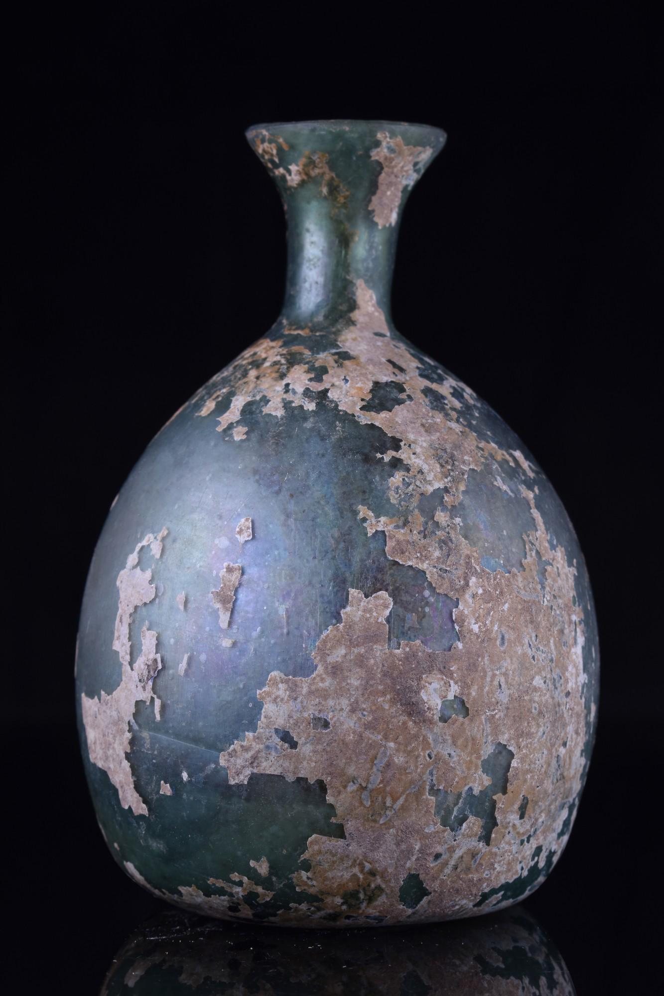 ANCIENT ROMAN GLASS FLASK: Ca. 100-300 AD.A beautiful green, free-blown glass bottle featuring a squat body on a concave base, a short cylindrical neck, and an everted rim. Some weathering and incrustation occur throughout the 
