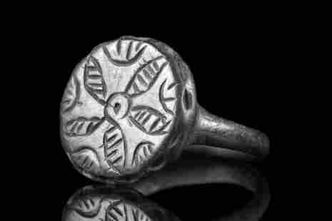 BYZANTINE BRONZE RING WITH FLORAL PATTERN: Ca. 500-600 AD. A beautiful bronze ring with a smooth, circular band supporting a large bezel decorated with a five-petaled floral design. Further incised decoration runs around the perimeter of the b
