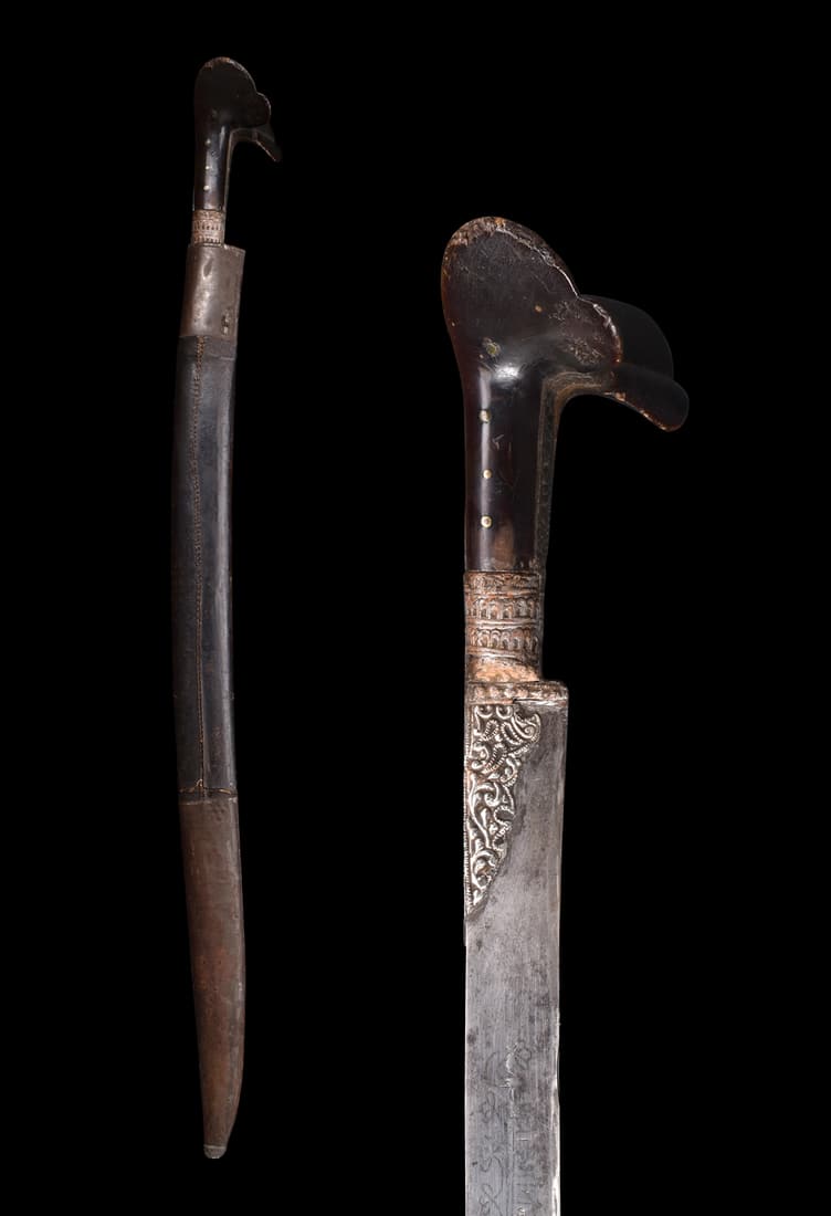 OTTOMAN YATAGAN SWORD WITH SILVER INLAYS AND A SCABBARD: Ca. early 19th century AD.A Yatagan sword with its hilt crafted from silver exhibits a distinctive eared shape. It is adorned with intricate repousse and engraving, showcasing the skilled artistry of 