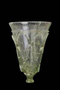 RARE MIGRATION PERIOD CUT GLASS BEAKER: Ca. 500-700 AD. A pale green cut glass beaker of a conical shape, standing on a small, rounded foot, providing balance and stability. The exterior walls are adorned with exquisite petal-shaped