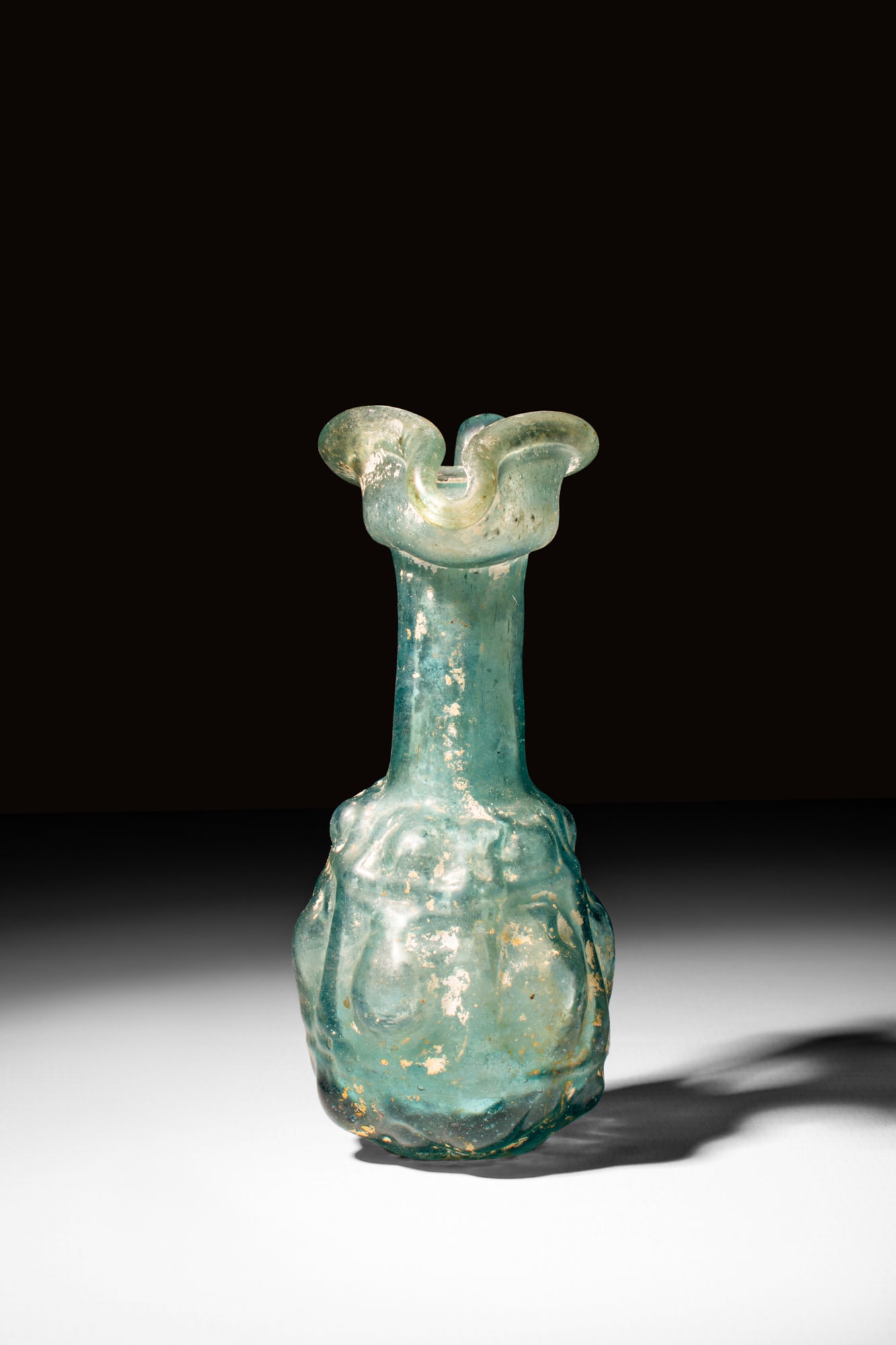 ROMAN GLASS OINOCHOE WITH TREFOIL SPOUT: Ca. 3rd - 4th century AD. A glass oinochoe featuring a rounded lower body with moulded decoration, tubular neck with trefoil spout and an elegantly arched handle connecting the rim with a shoulder. Th