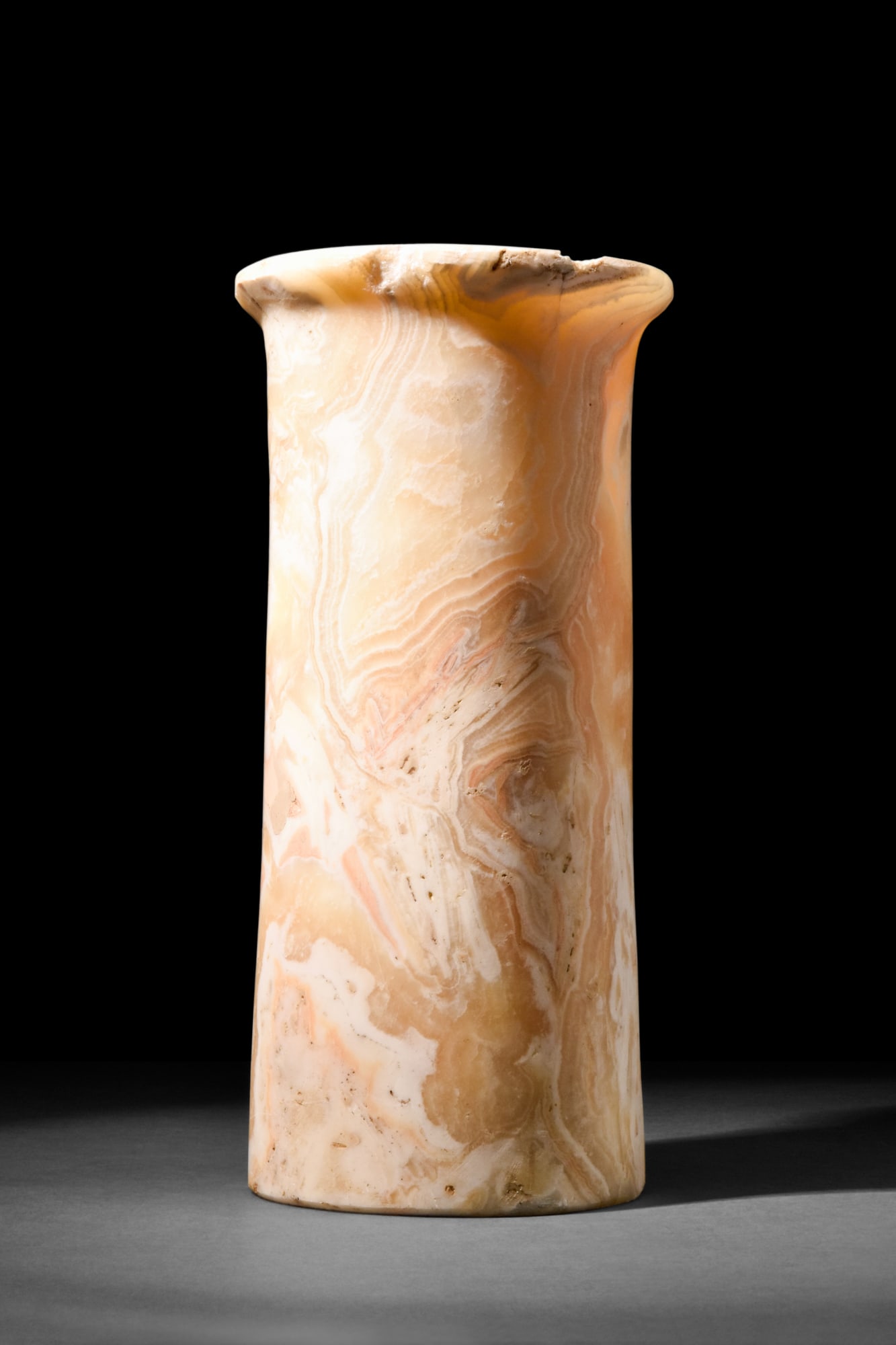 EGYPTIAN ALABASTER TALL VESSEL: The Third Dynasty, Ca. 2650-2575 BC.A tall cylindrical vessel with a rounded and flaring rim. It rises from a flat circular base and features a hollow interior. Crafted from alabaster, this vessel sho