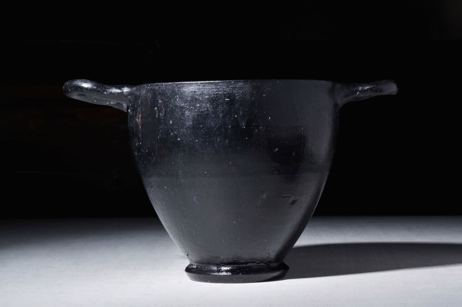 ATTIC BLACK-GLAZED SKYPHOS: Ca. 350 BC.A black slip skyphos featuring deep, curved sides, resting on a sturdy ring foot. The vessel is fitted with two equally spaced loop handles, positioned on either side of the cup. The skypho