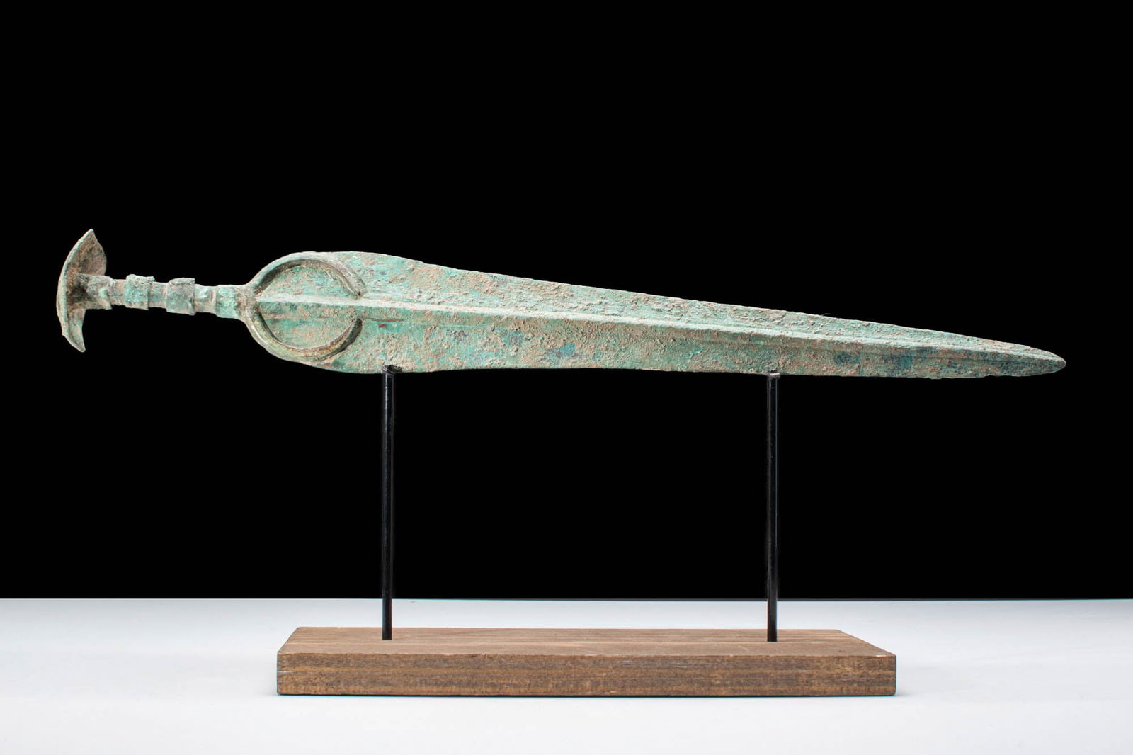 ANCIENT BRONZE SWORD WITH CRESCENTIC POMMEL: Western Asiatic/Aegan, Ca. 1200 - 700 BC. A bronze sword with a long tapering blade, featuring a raised mid-rib and a curved guard for hand protection. The hilt has a round-section shank, and the swor