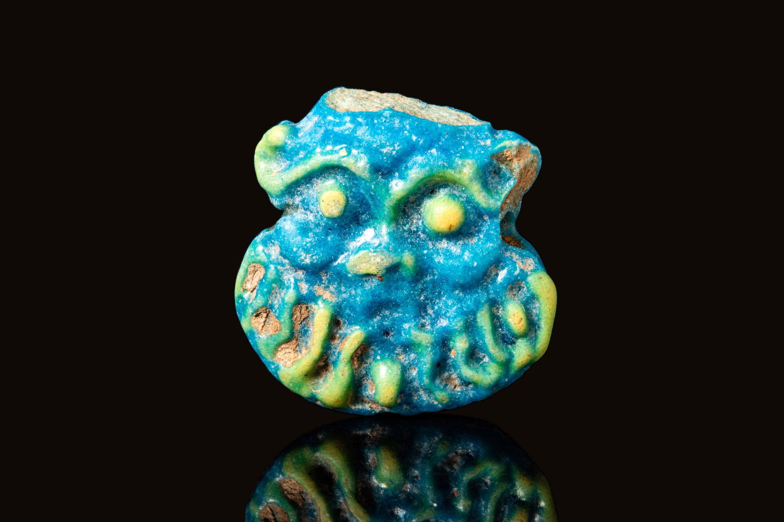 EGYPTIAN FAIENCE AMULET OF BES: New Kingdom, Ca. 1550 - 1069 BCAn Egyptian mould-formed faience amulet of the deity Bes with a long beard. Bes's head is represented frontally with yellow details of the eyes, the eyebrows, and the be