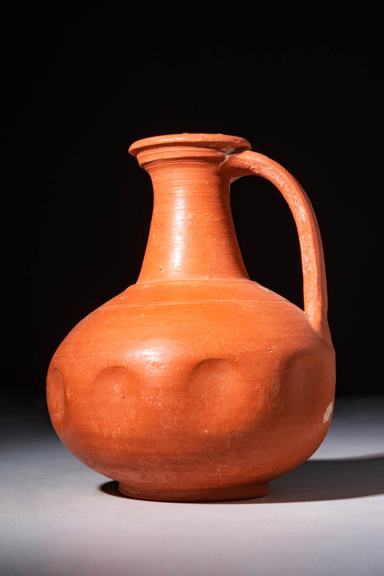 ROMAN TERRA SIGILLATA POTTERY WINE FLAGON: Ca. AD 100 - 300A Roman terra sigillata redware flagon with an apple-shaped body and a squat foot underneath. It has a narrow neck and flat rim. Projecting from the shoulder is a handle that is patter