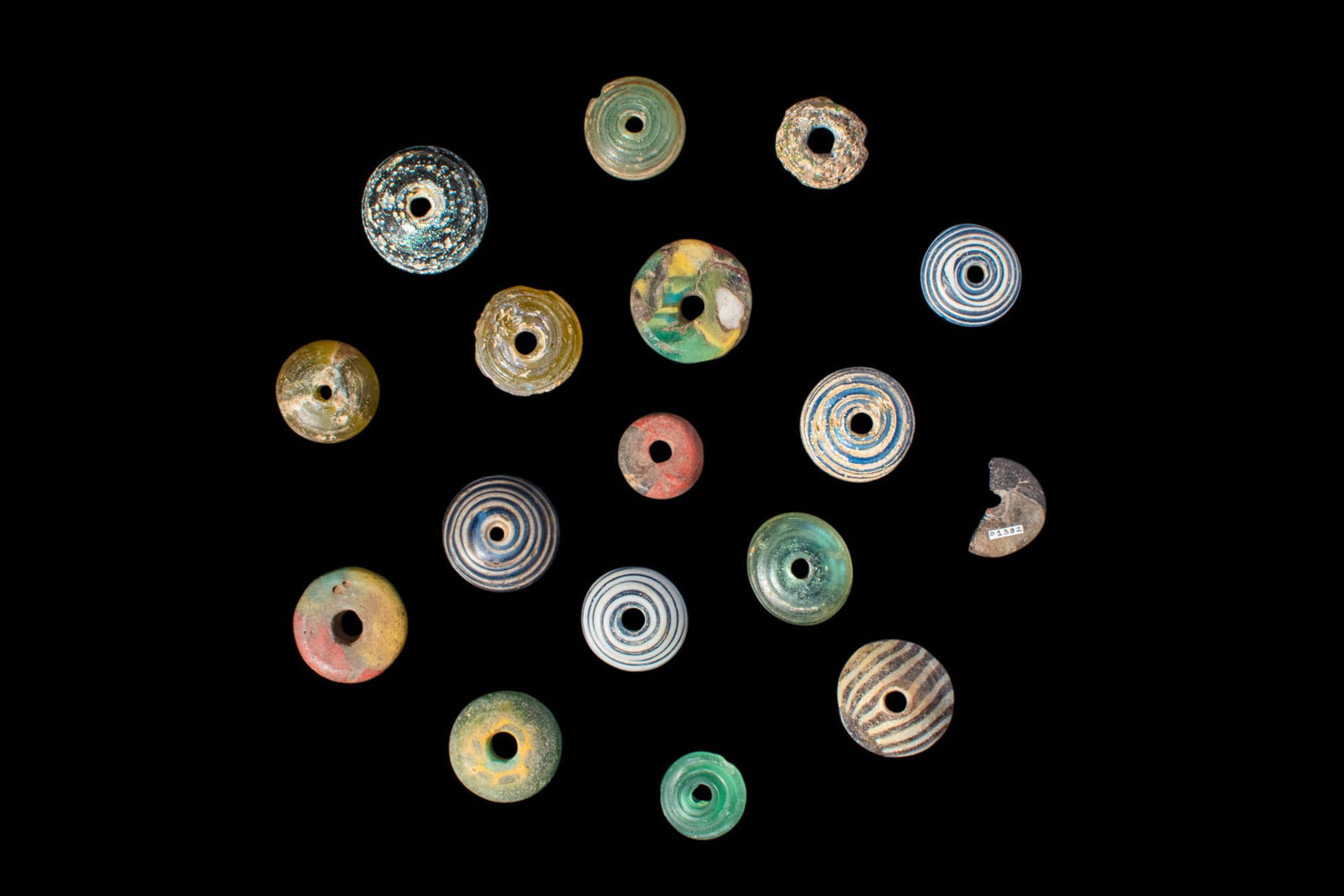 COLLECTION OF ROMANO - EGYPTIAN GLASS BEADS: Ca. 30 BC - AD 200.A collection of 17 glass beads, shaped as circular, flat discs with slightly domed upper sides. Each piece is vertically perforated at the centre, exhibiting variations in hues, siz