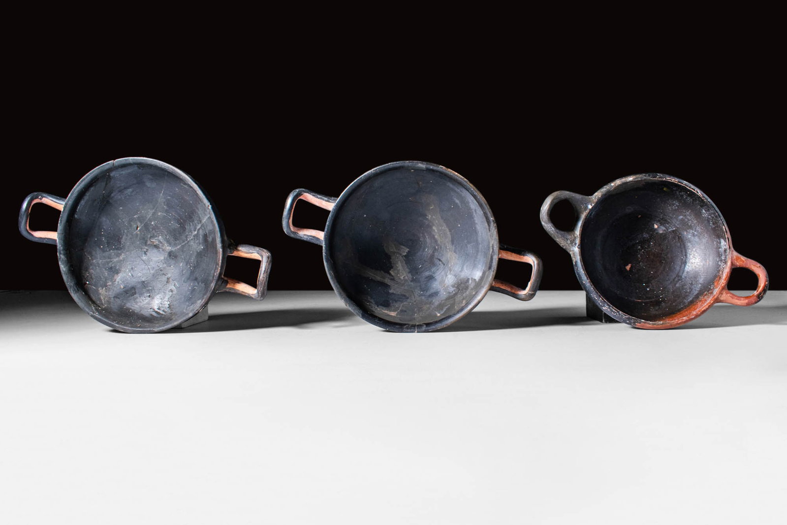 SET OF GREEK BLACK GLAZE KYLIKES: Ca. 400 - 300 BC. A set of three elegant blackware kylikes. The first kylix, on the left, has a carinated and deep body. The vessel has two horizontal handles and is seated on a high pedestal. The sec