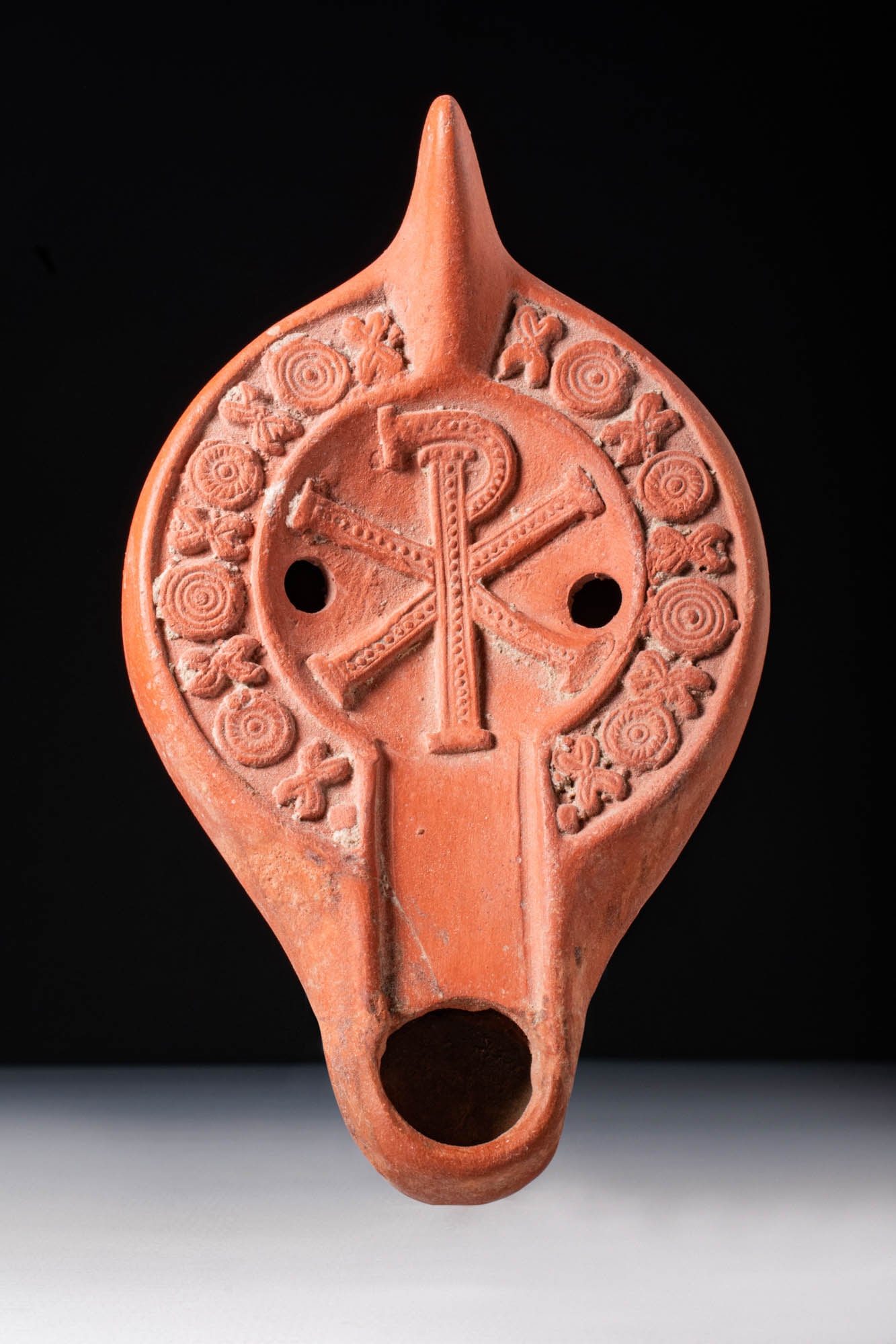BYZANTINE TERRACOTTA OIL LAMP WITH CHI-RHO: Ca. AD 400 - 500. A red terracotta oil lamp with a rounded body and a sunken upper face bordered by a ridge and channelled to a spout. The central discus displays a relief depiction of a Chi-Rho, an e