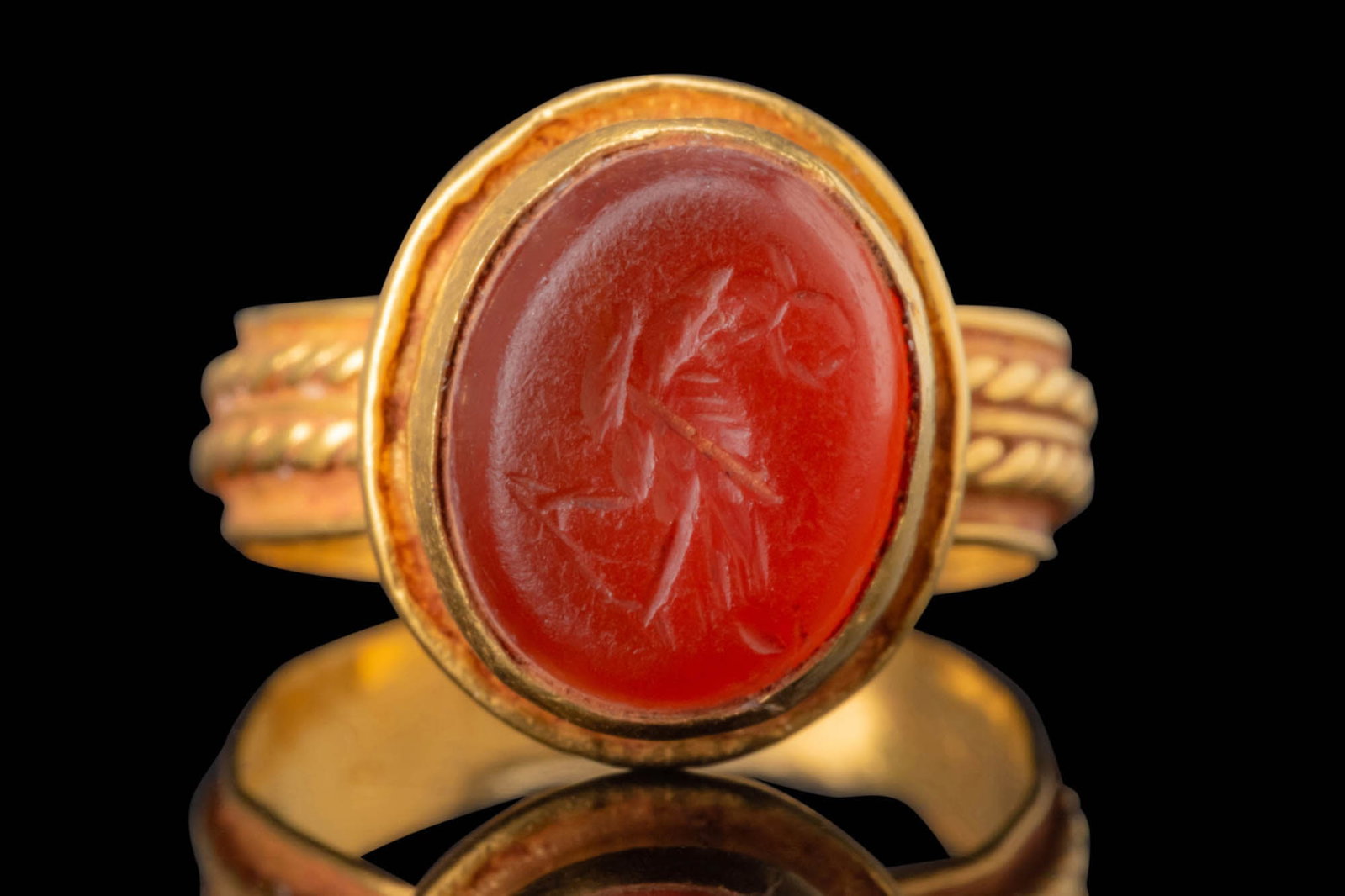 BYZANTINE CARNELIAN INTAGLIO WITH LEGIONARY EAGLE IN GOLD RING: Ca. AD 300 - 500. An oval carnelian intaglio engraved with a standing eagle grasping a wreath in its beak. The body displays incised lines forming the feathers, while the legs extend downward, ending 