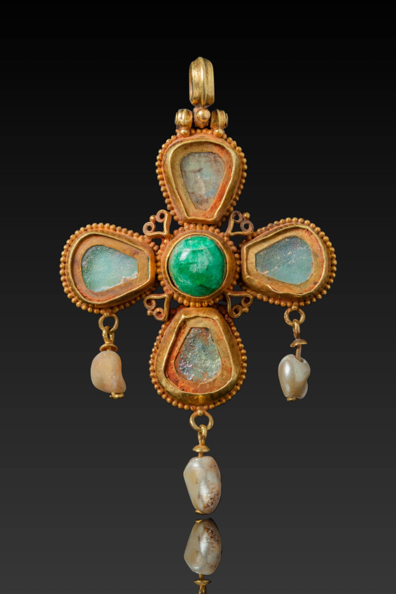 BYZANTINE HIGH KARAT GOLD CROSS PENDANT WITH CENTRAL EMERALD AND DANGLING PEARLS: Ca. AD 600. A gold cross pendant centred with a domed emerald set within a beaded border. Each arm is mounted with a drop-shaped glass insert framed in gold with a granulated outline. Openwork heart-s