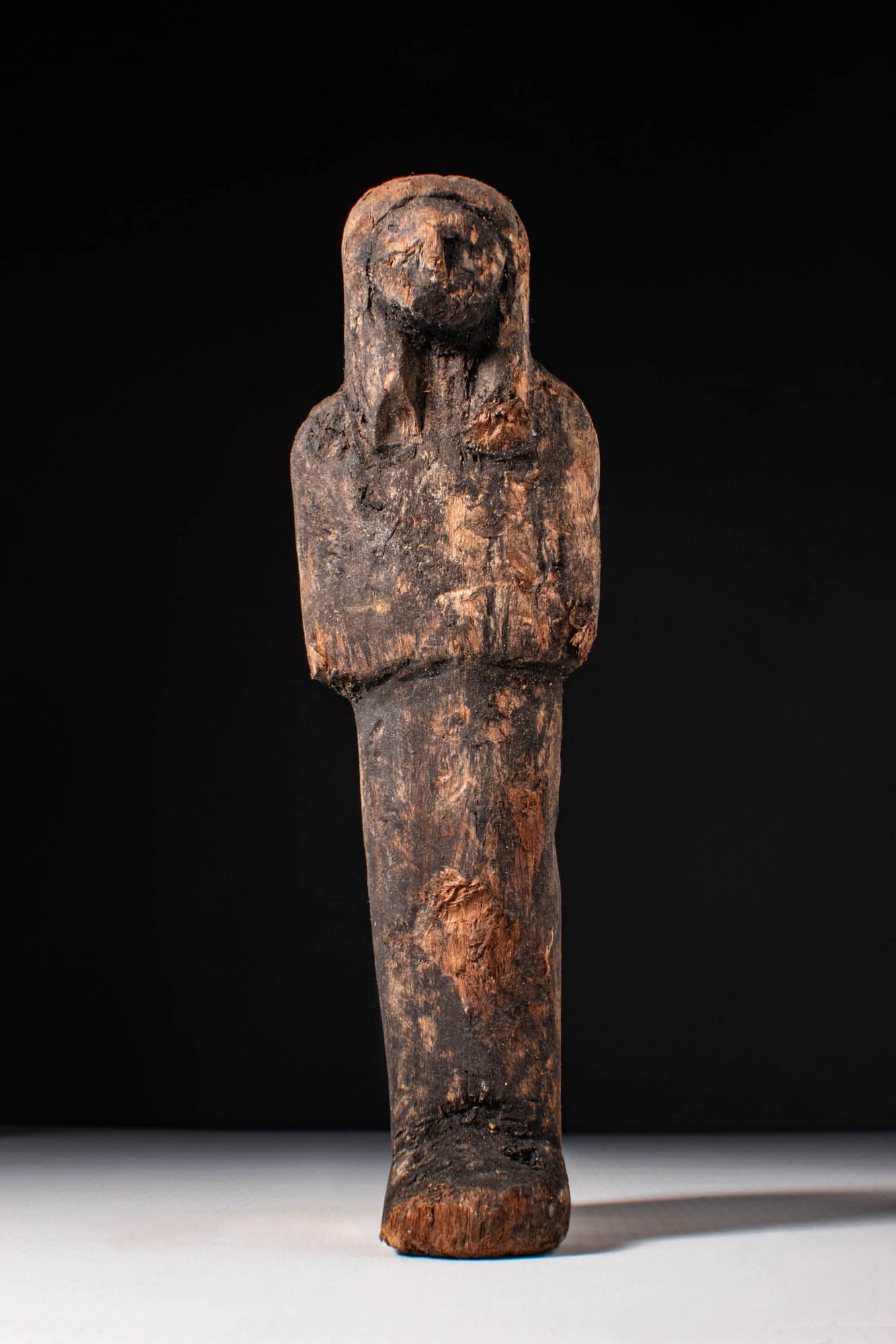 EGYPTIAN WOODEN USHABTI: New Kingdom, Ca. 1550 - 1069 BC. A wooden ushabti figure depicted standing with hands placed on the chest and wearing a tripartite wig. Its side lappets fall over the shoulders and extend down the upp