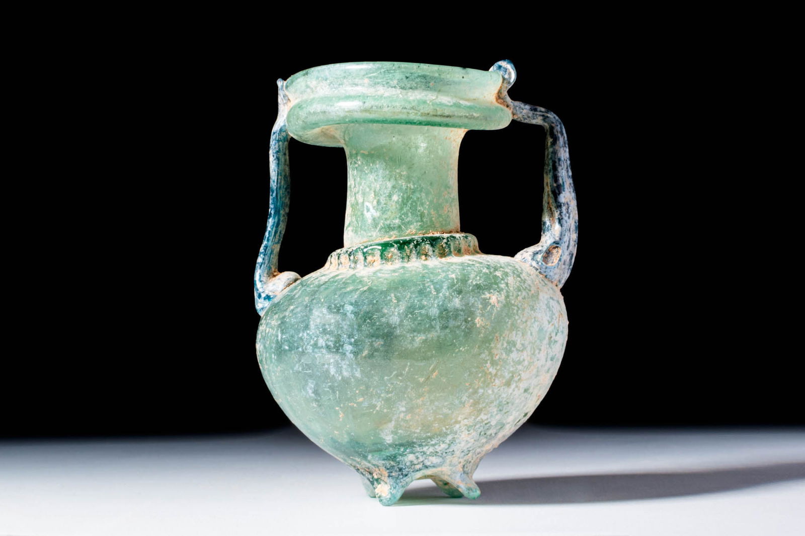 ROMAN GLASS AMPHORISKOS: Ca. AD 300 - 500. A translucent pale green glass amphoriskos with a globular body tapering to a pointed base with four rounded feet. The cylindrical neck widens into a thick, rounded rim with an outwa